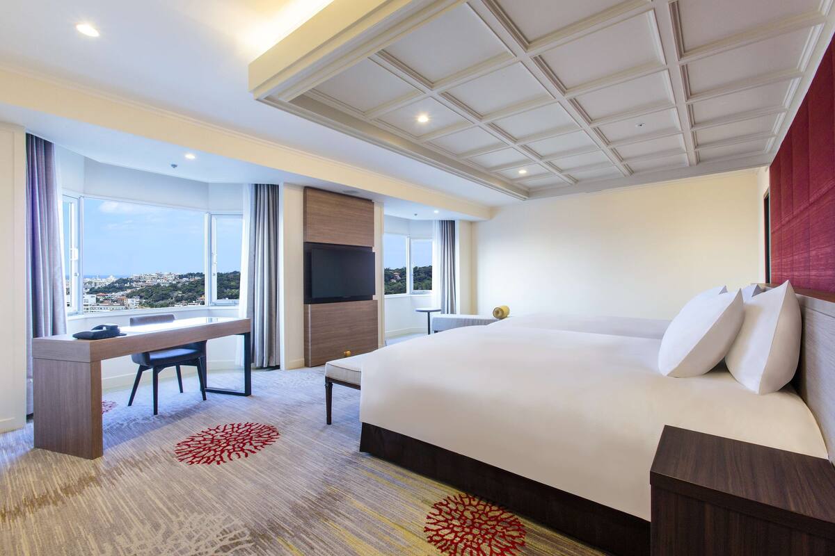 A hotel room with a large bed, a desk with a chair, a TV, and a view of the city.