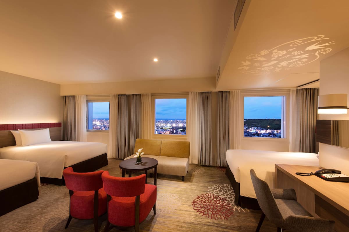 A hotel room with two beds, a sofa, a desk, and chairs. There are large windows with a city view.
