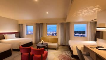 A hotel room with two beds, a sofa, a desk, and chairs. There are large windows with a city view.