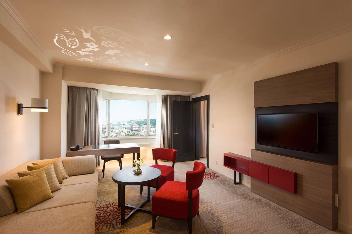 A modern hotel room with a sofa, red chairs, a dining table, a TV mounted on the wall, and a large window offering a city view.