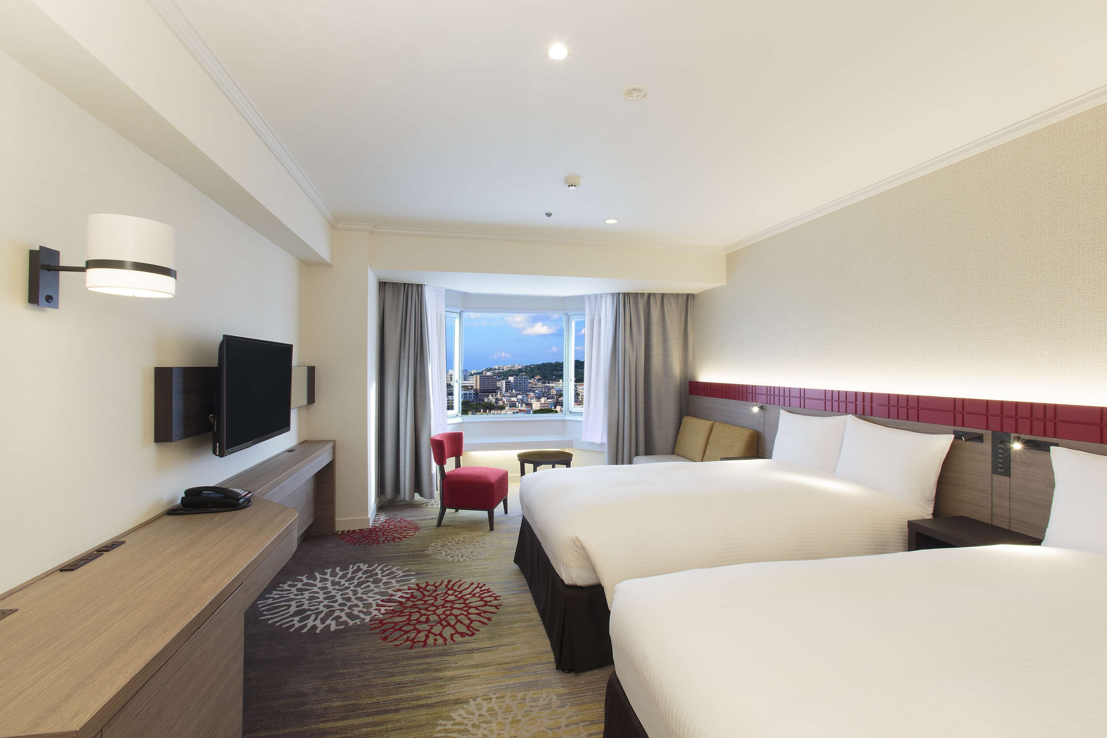 A hotel room with two beds, a TV, a desk, and a view of a cityscape.