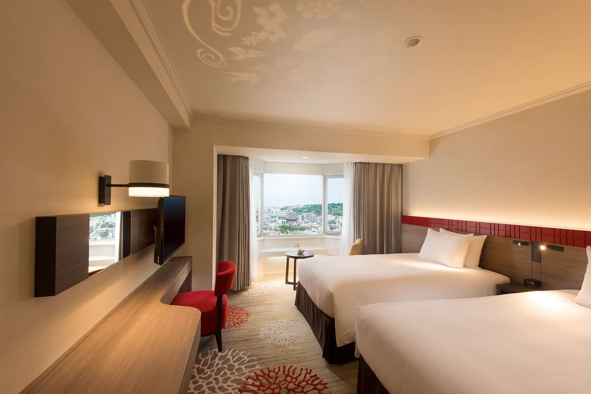 A hotel room with two beds, a TV, a small table, and a view of the city.