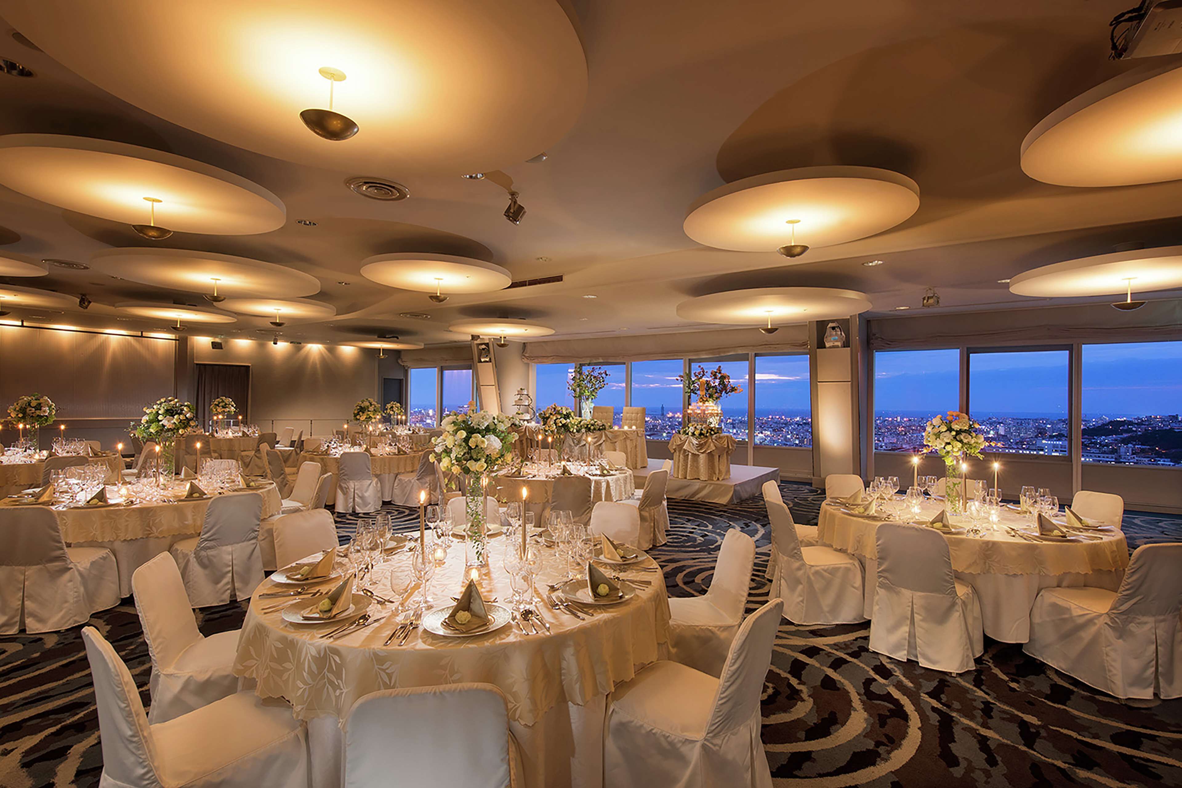 A banquet hall with round tables set for a formal event, large windows with a city view, and a modern ceiling design.