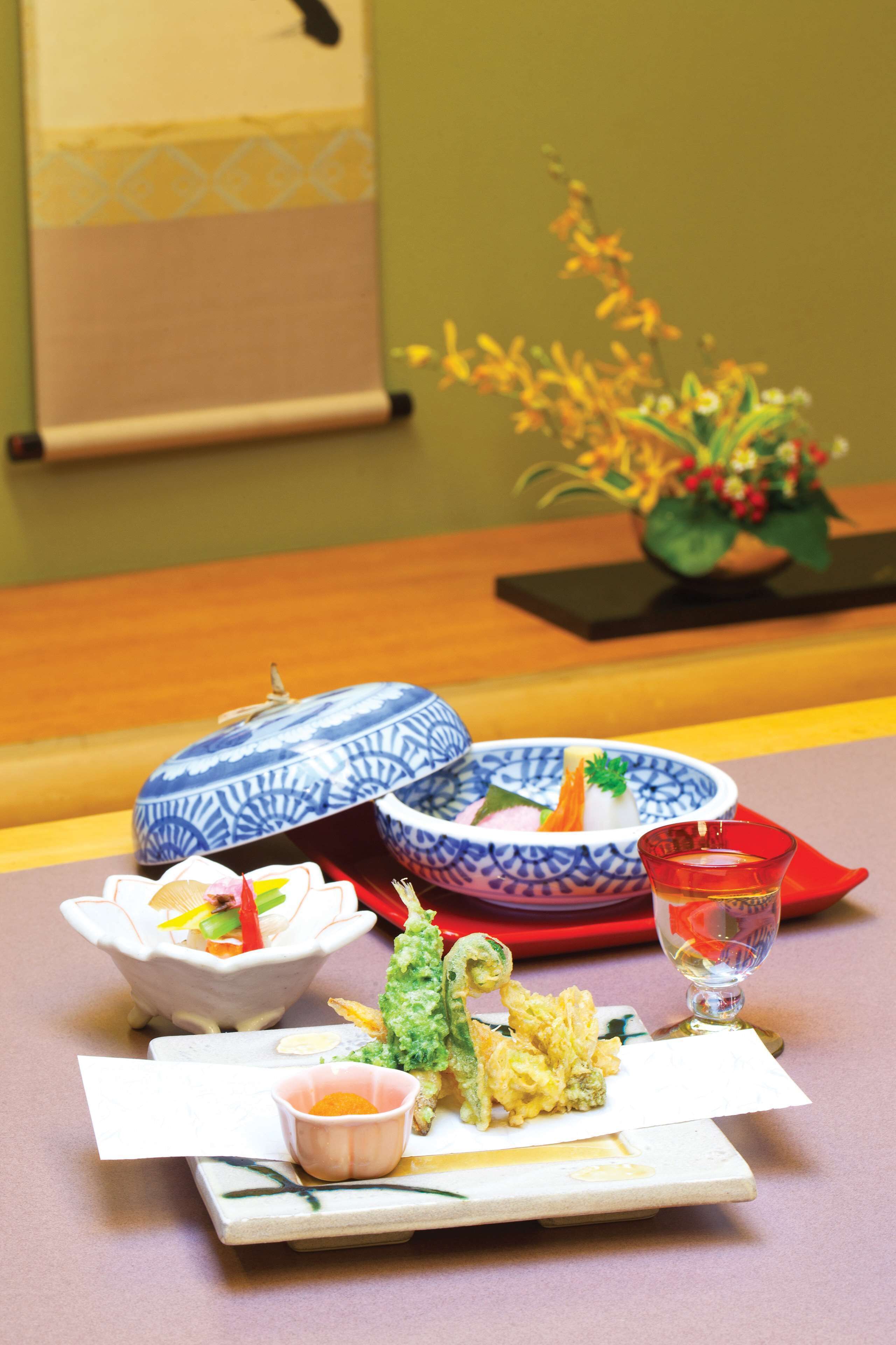 A table set with a blue and white patterned bowl, a white bowl with a green garnish, a red napkin, a glass of red liquid, and a small dish with a yellow garnish.