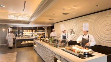 A modern restaurant with a central counter where chefs are preparing food. There is a display case with various items and a seating area with tables and chairs.