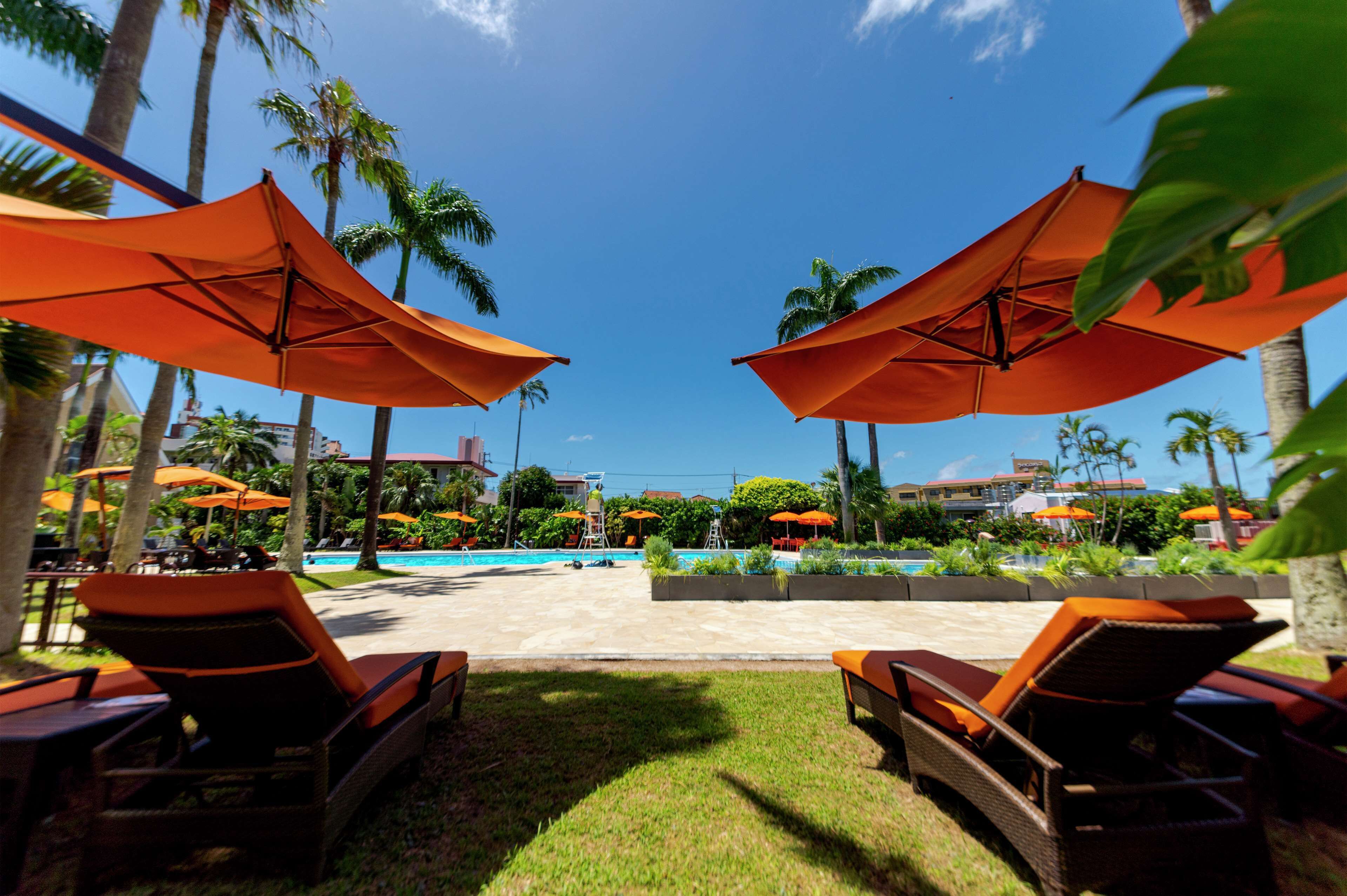 A poolside area with lounge chairs and orange umbrellas, surrounded by palm trees and a clear blue sky.