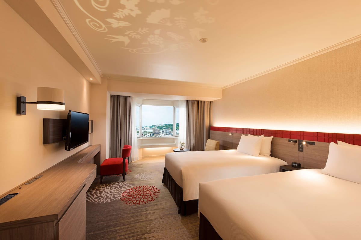 A hotel room with two beds, a TV, a desk, and a view of the city.