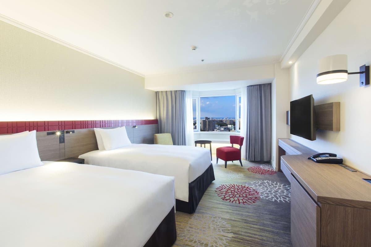 A hotel room with two beds, a desk, a chair, a TV, and a window with a city view.
