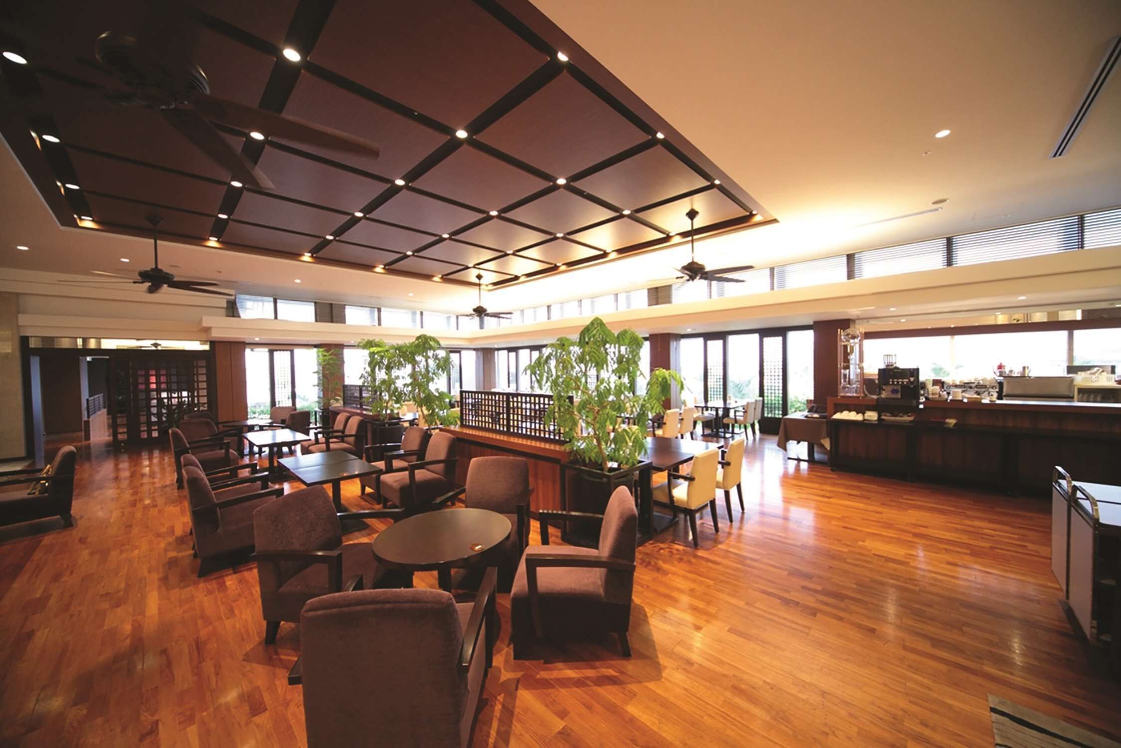 Un espacio interior amplio con suelo de madera, varios conjuntos de asientos, ventiladores de techo y grandes ventanales que dejan entrar luz natural.