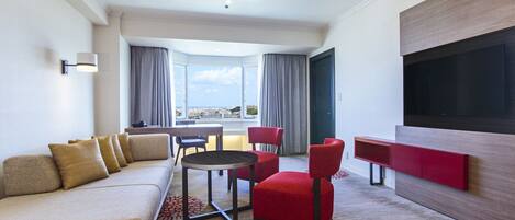 Habitación de hotel con sofá, sillas, una mesita, televisor y vistas a la ciudad.