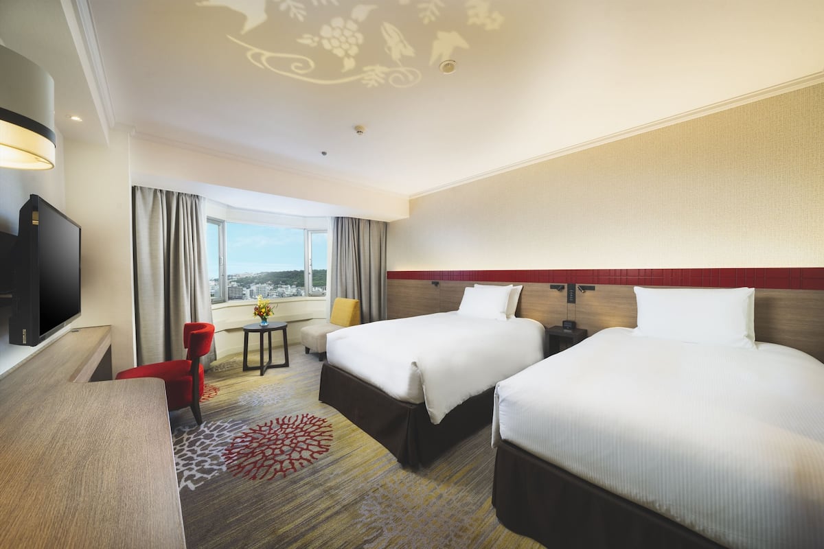 A hotel room with two beds, a TV, a desk, a chair, and a view of the city.