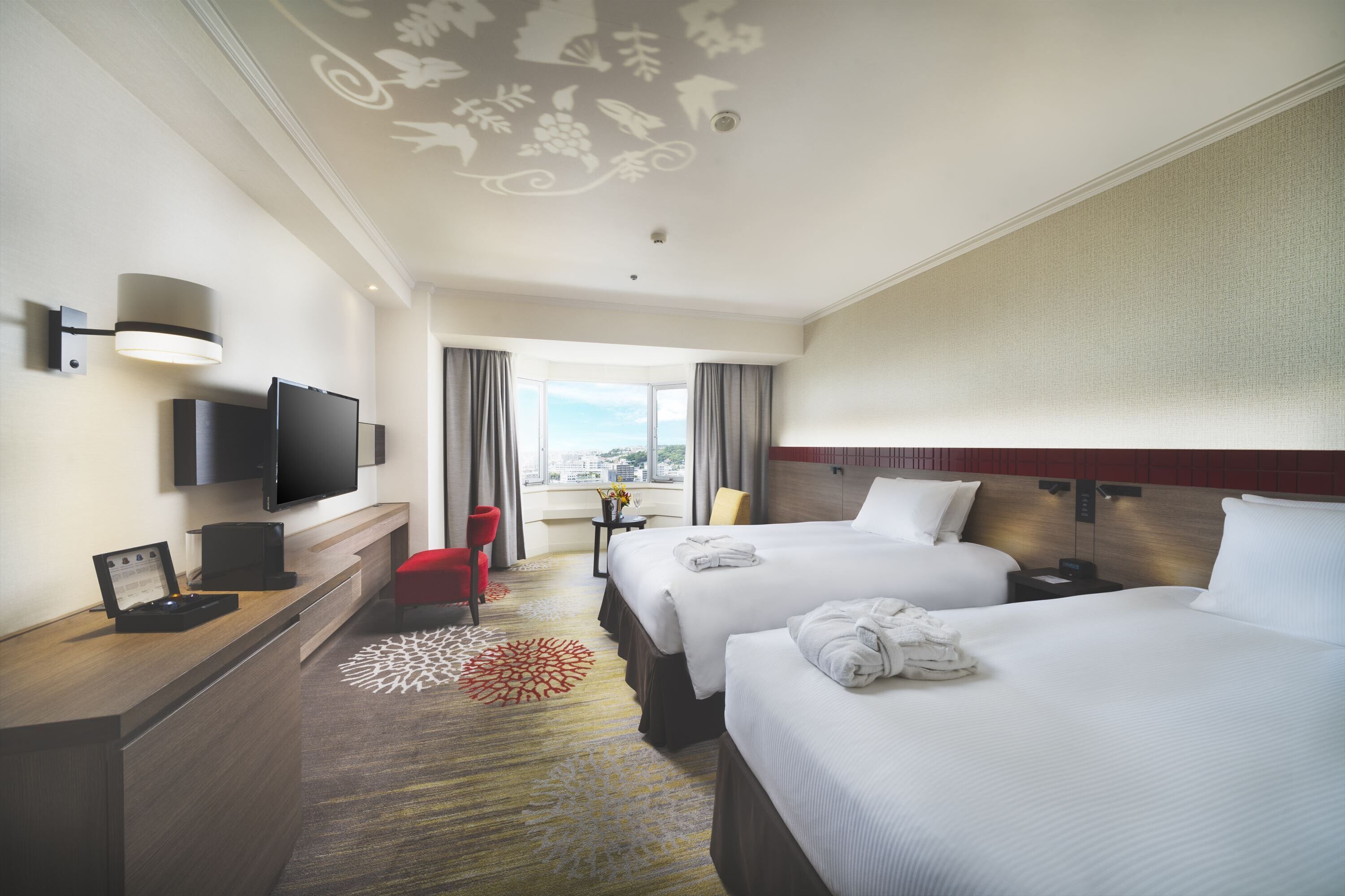 Habitación de hotel con dos camas, un escritorio, un televisor de pantalla plana, una silla roja y vistas a la ciudad a través de amplios ventanales.