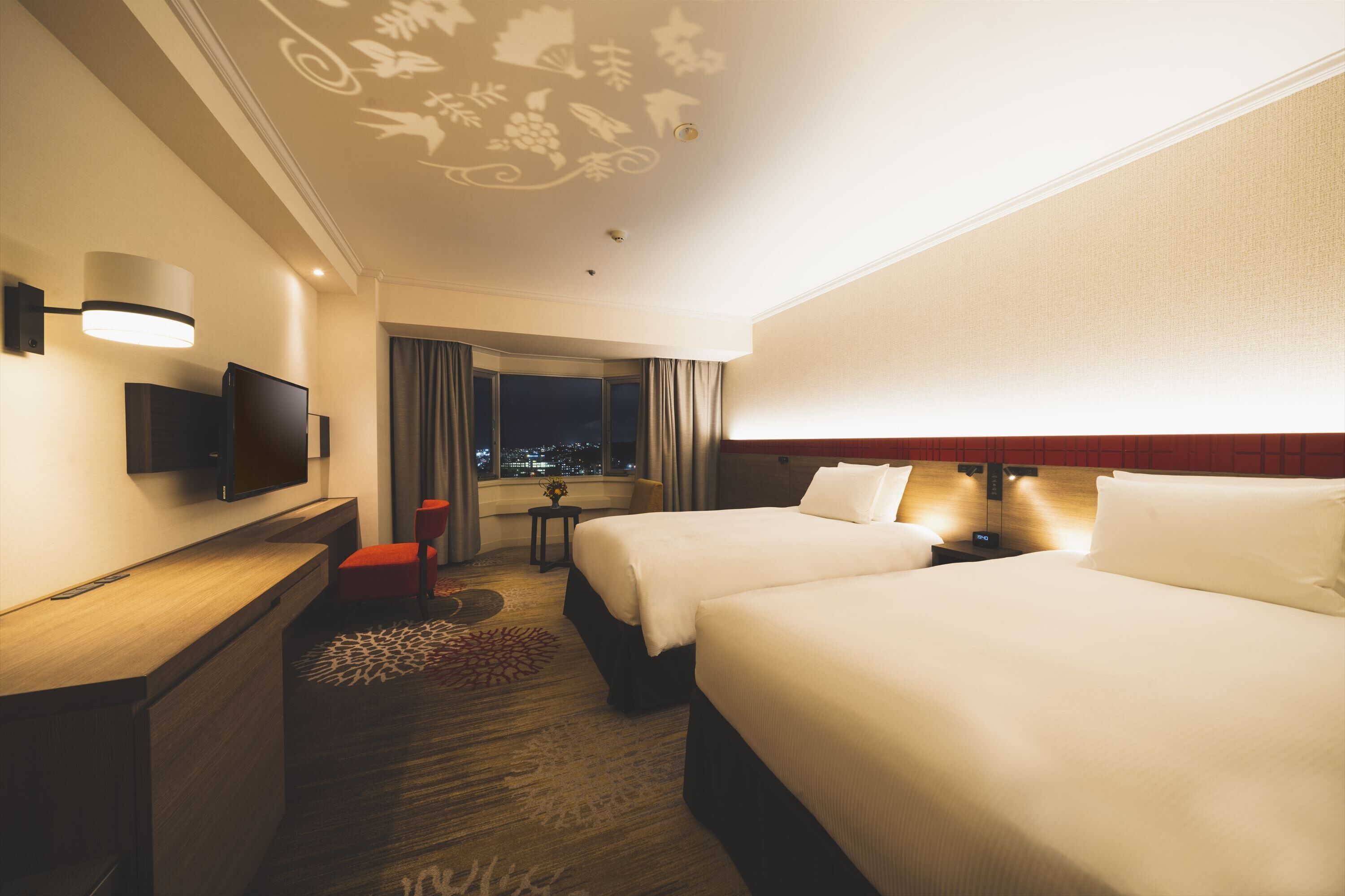 A hotel room with two beds, a desk with a flat-screen TV, a small table with a vase, and a view of the city at night.