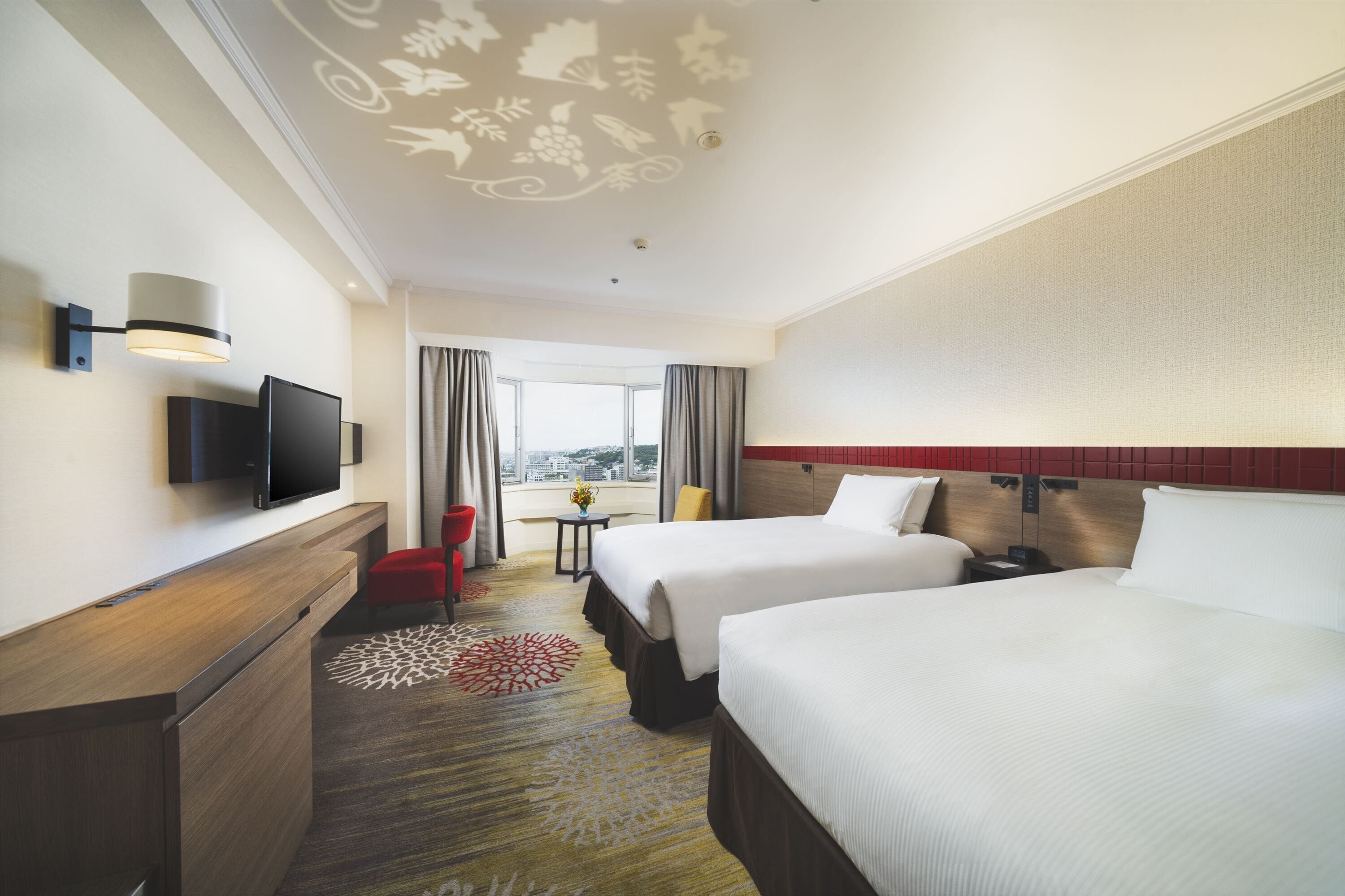 Une chambre d’hôtel avec deux lits, un bureau doté d’un téléviseur à écran plat, une chaise rouge, une petite table avec un vase, et une vue sur la ville grâce à de grandes fenêtres.