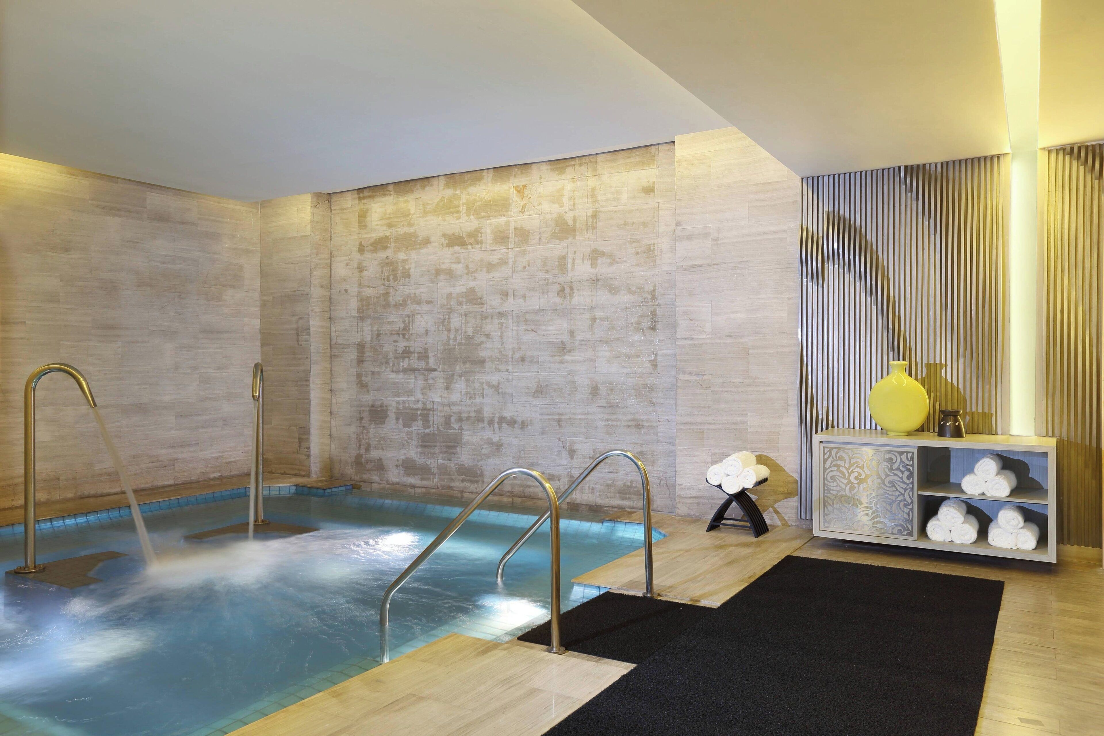 Un moderno espacio de spa con un jacuzzi, barandales de metal, suelo de madera y un mueble decorativo con toallas.