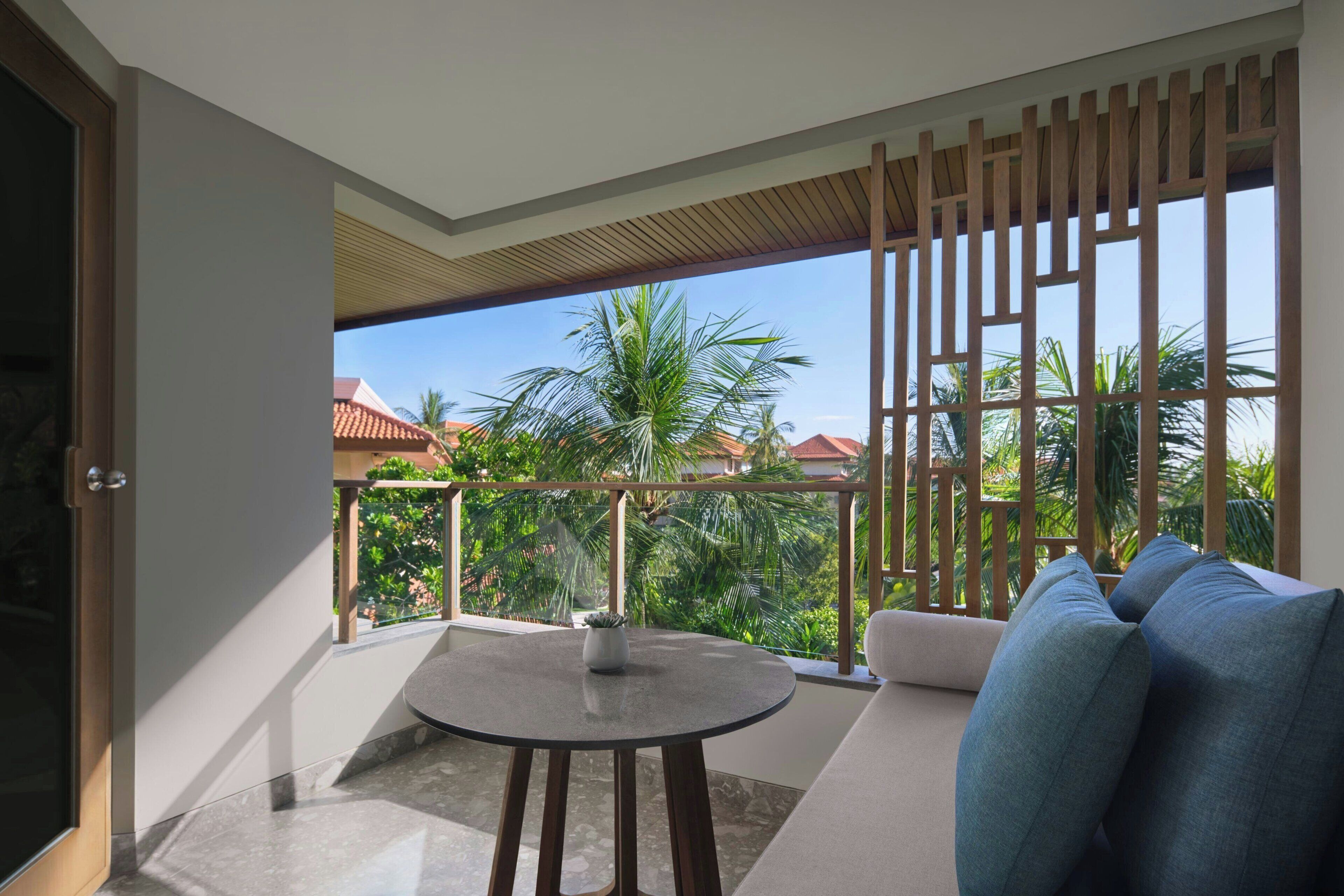 A balcony with a round table, a sofa with cushions, and a view of palm trees and buildings.