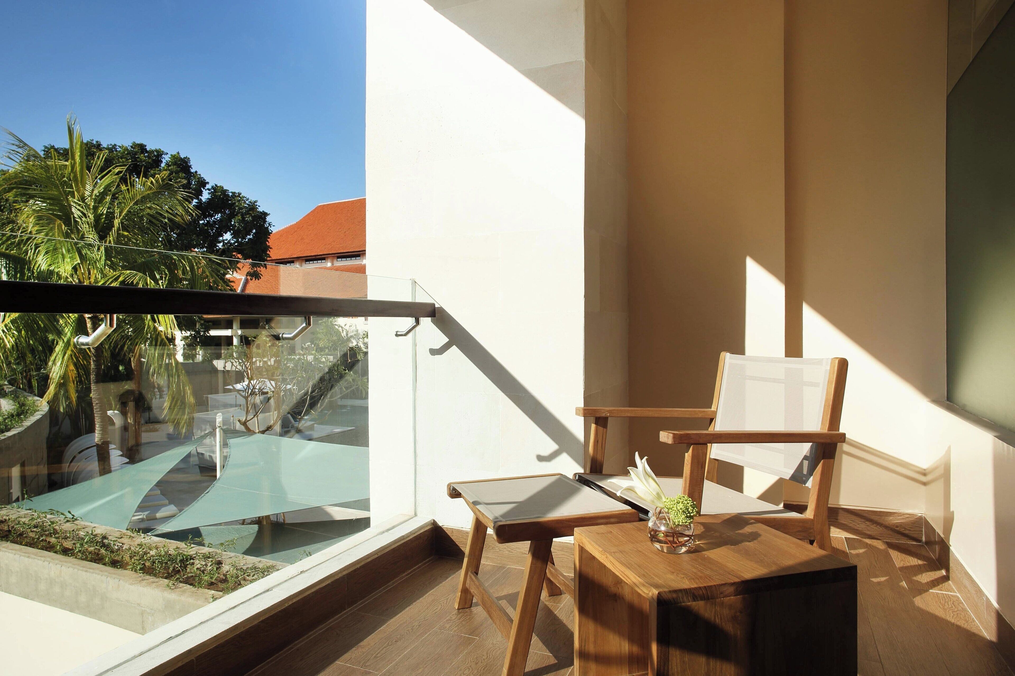 A balcony with a glass barrier, wooden furniture, and a view of a building and palm trees.