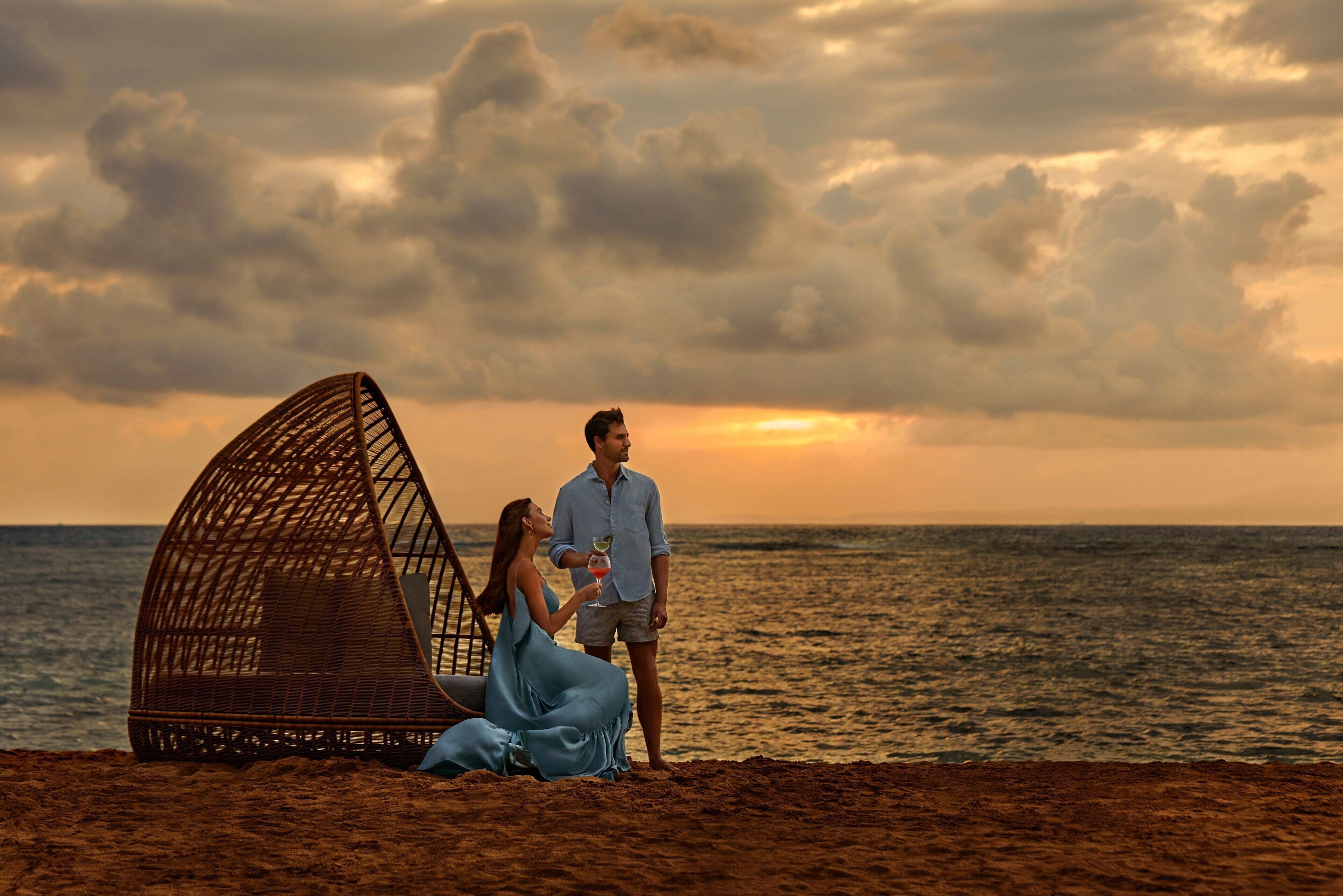 A beach scene with a large, open, woven structure and a couple sitting on the sand.