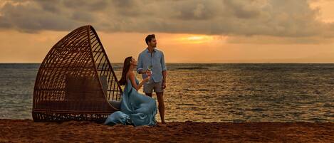 A beach scene with a large, open, woven structure and a couple sitting on the sand.