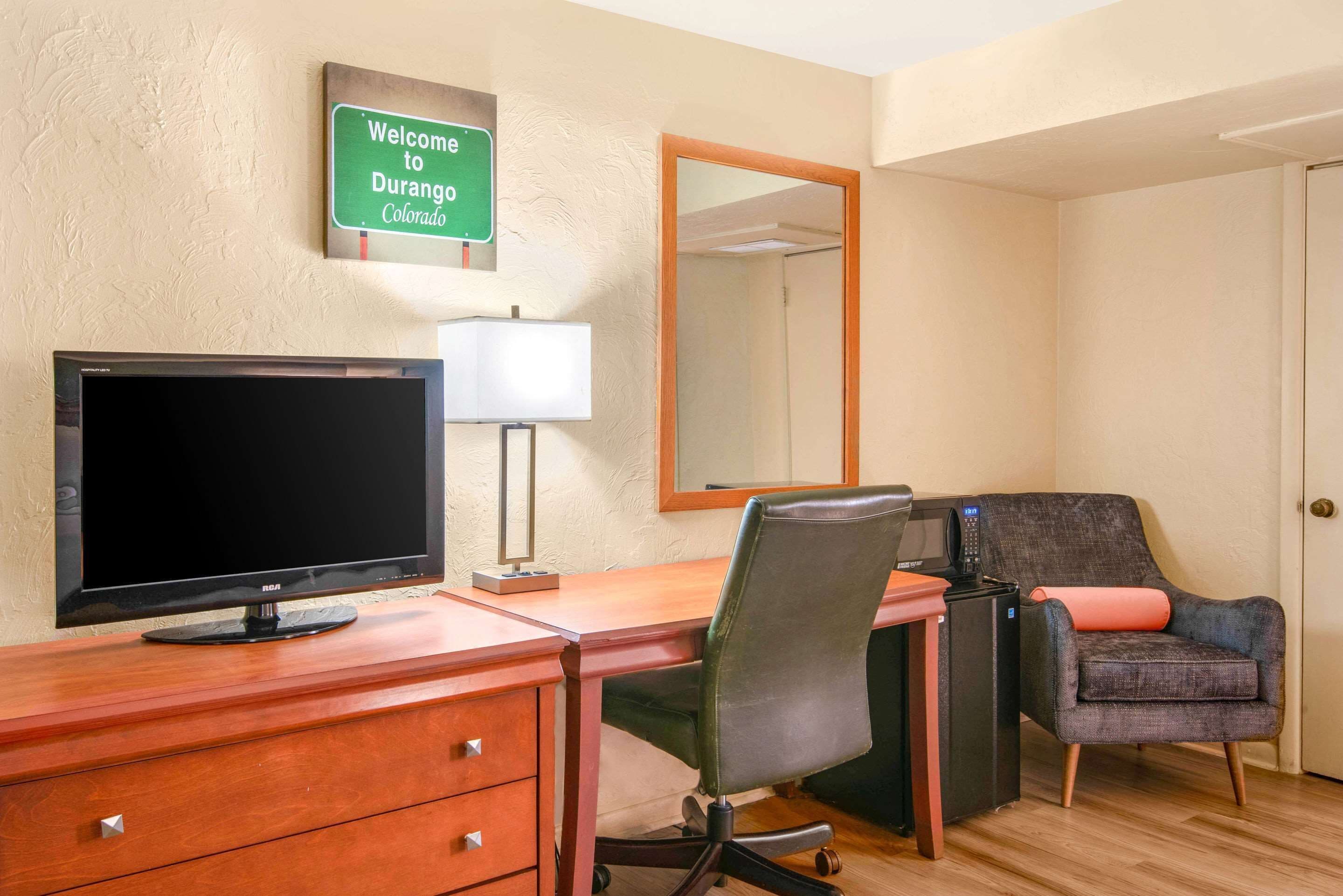 Une chambre d’hôtel avec un bureau en bois, une télévision, une chaise, un miroir et une pancarte de bienvenue.