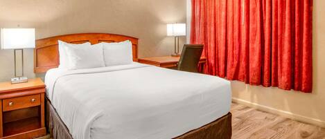 A hotel room with a large bed, wooden headboard, two bedside tables with lamps, a chair, and a red curtain.