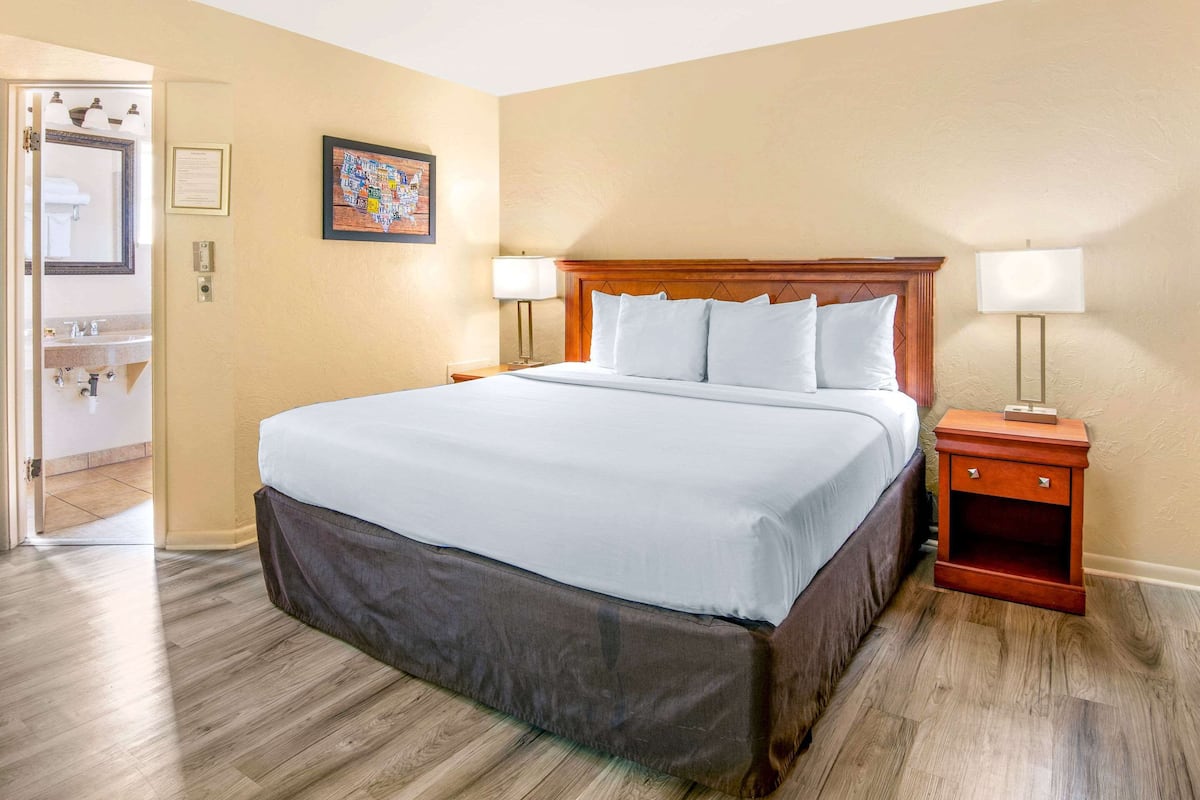 A hotel room with a large bed, wooden headboard, two bedside tables with lamps, a framed picture on the wall, and a bathroom visible through an open door.