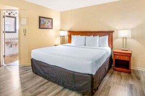 Quarto de hotel com uma cama grande, cabeceira de madeira, duas mesas de cabeceira com luminárias, um quadro emoldurado na parede e um banheiro visível por uma porta aberta.