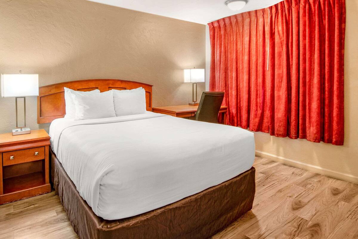 A hotel room with a large bed, wooden headboard, two bedside tables with lamps, a chair, and a red curtain.