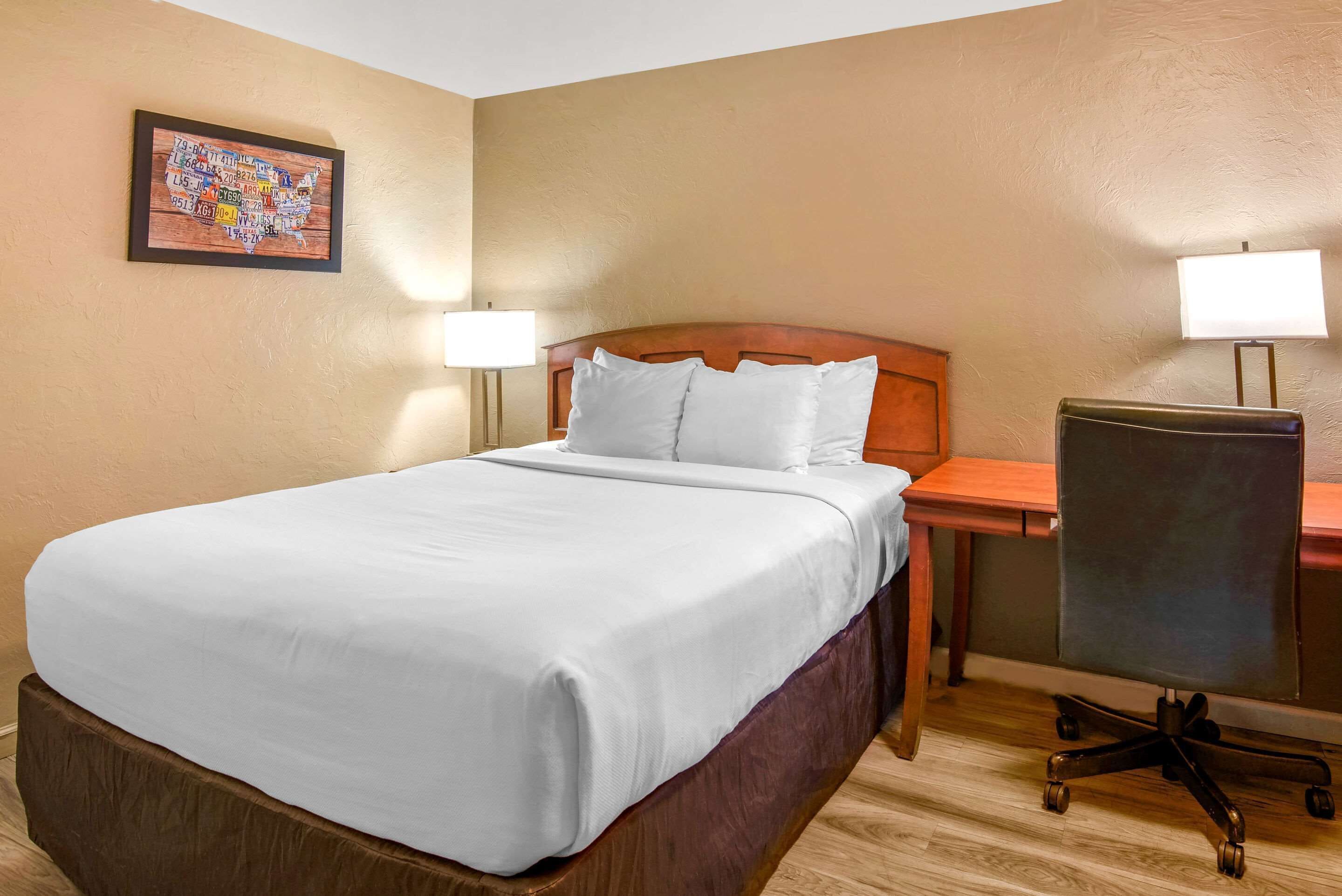 A hotel room with a bed, a desk, a chair, and a wall-mounted lamp.