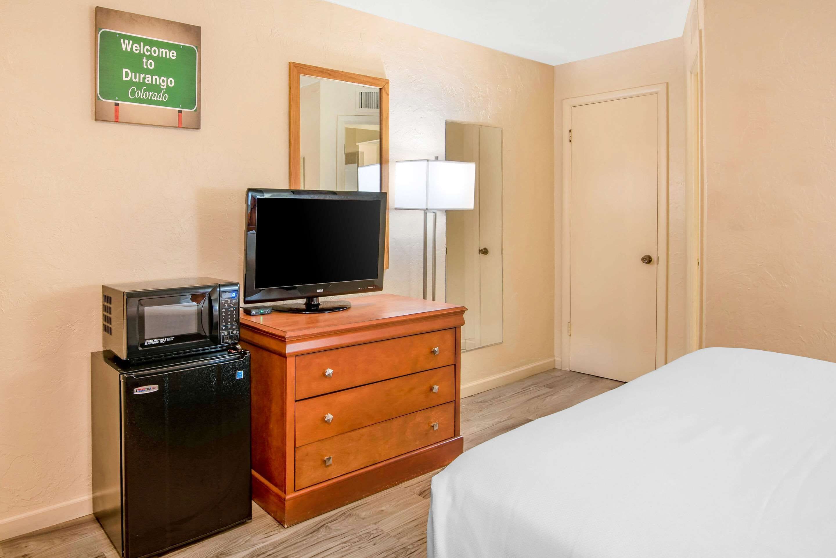 A hotel room with a bed, a wooden dresser with a television and a microwave, a door, and a sign on the wall.