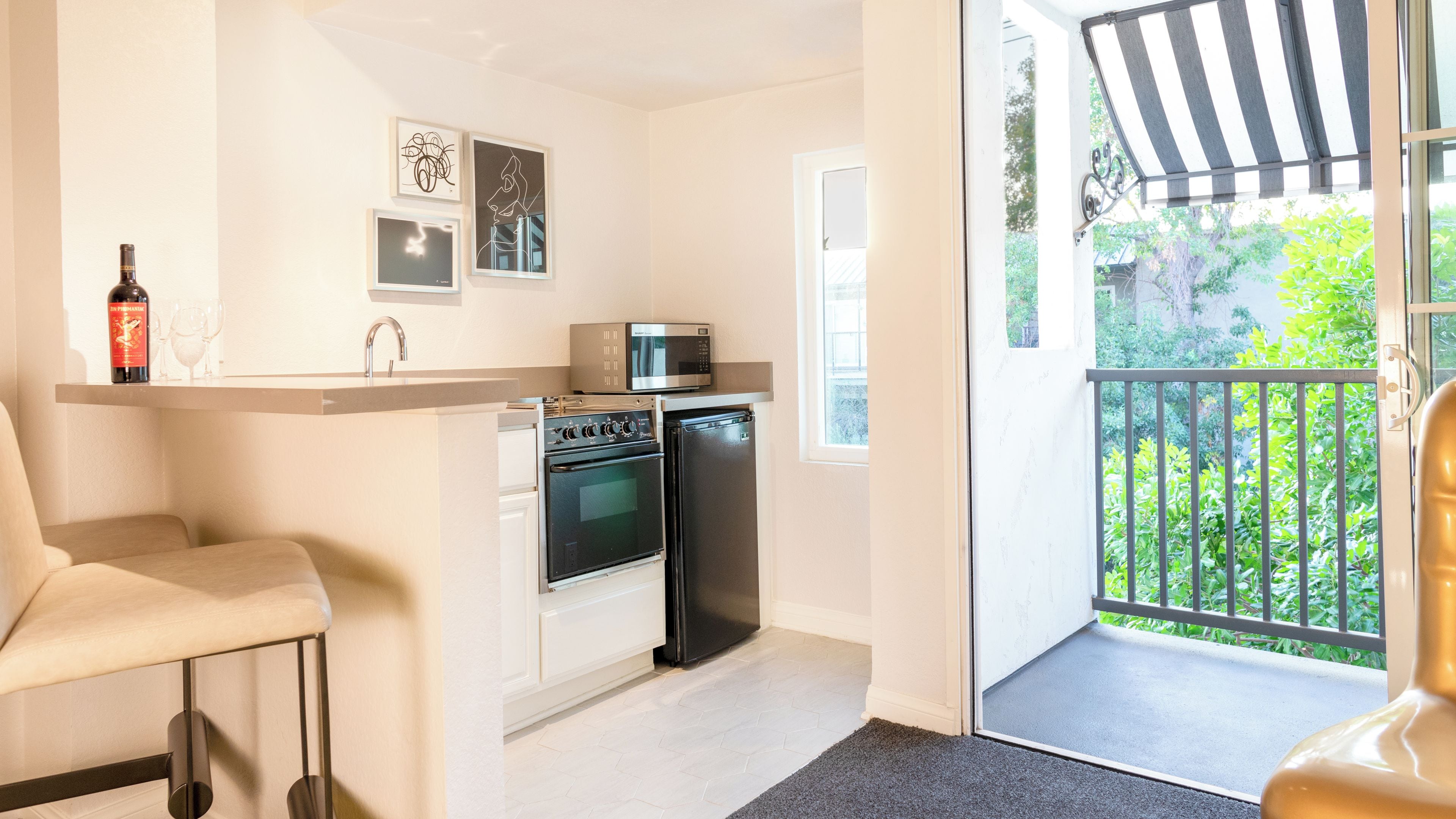 A compact kitchen with a microwave, oven, and refrigerator, a dining area with a chair, and a balcony with a view of greenery.
