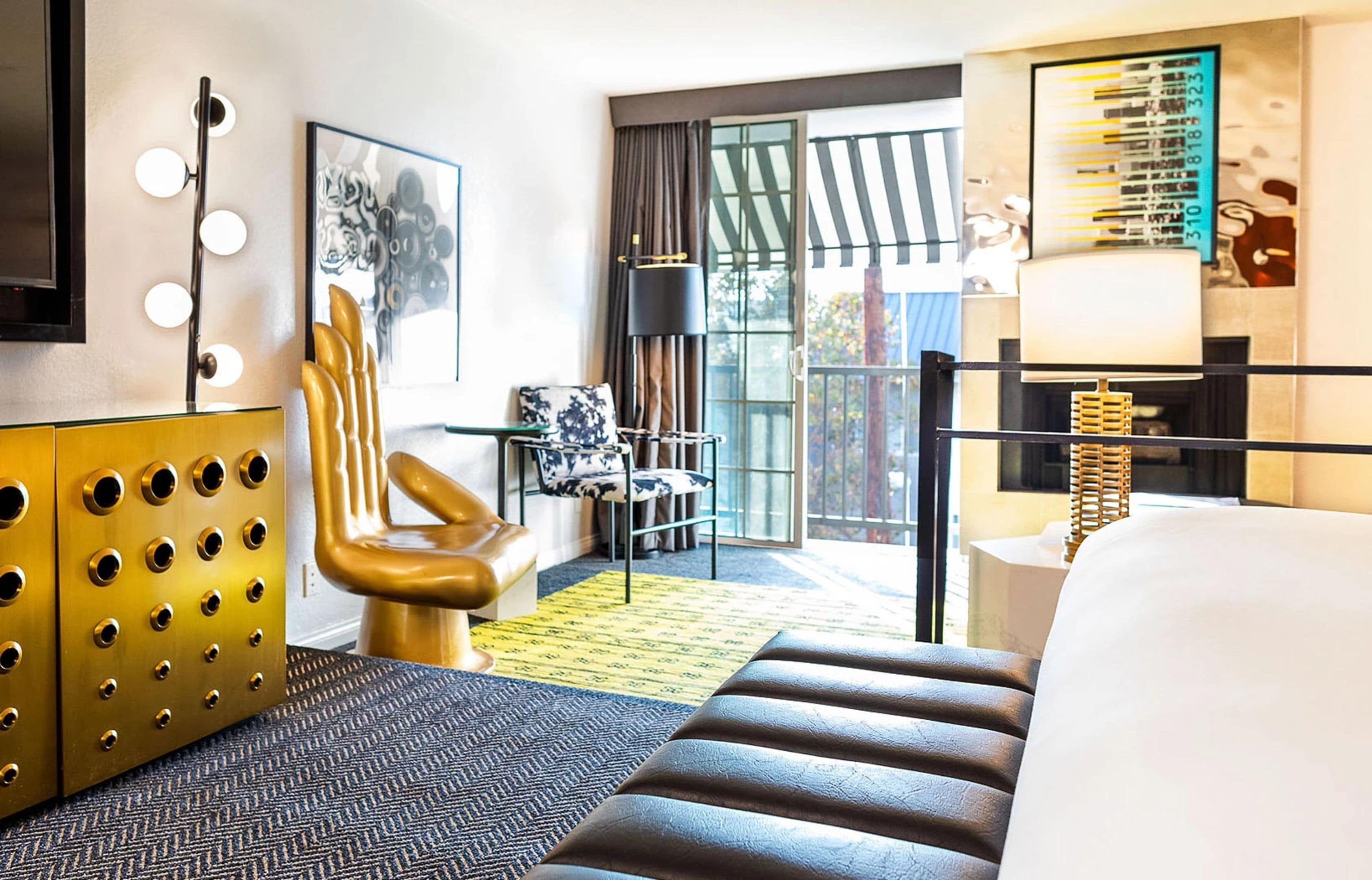 A hotel room with a large bed, a golden armchair, a small table, a floor lamp, and a large window with curtains.