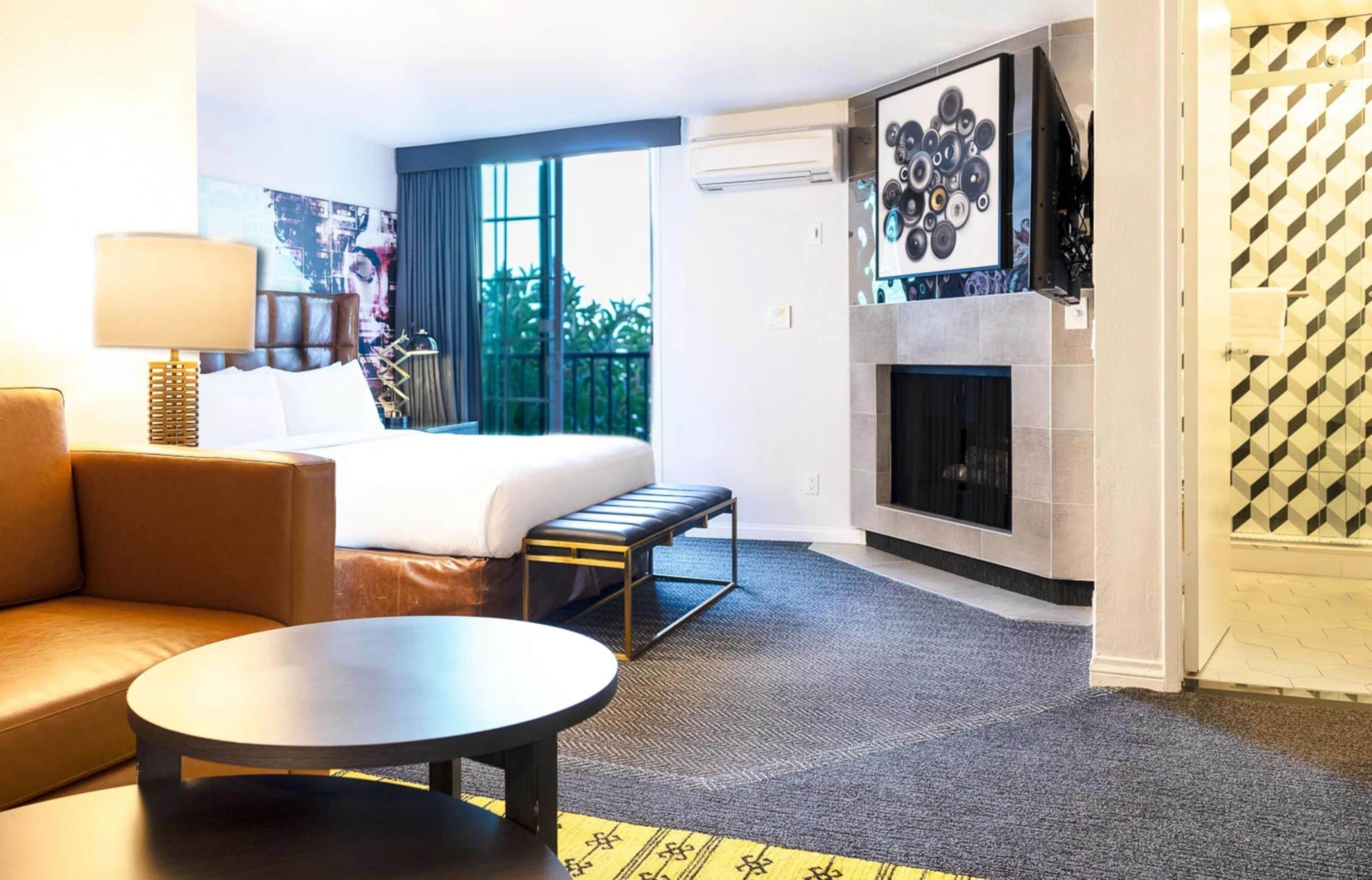 A hotel room with a bed, a sofa, a round table, a fireplace, and a large window with a view of greenery.