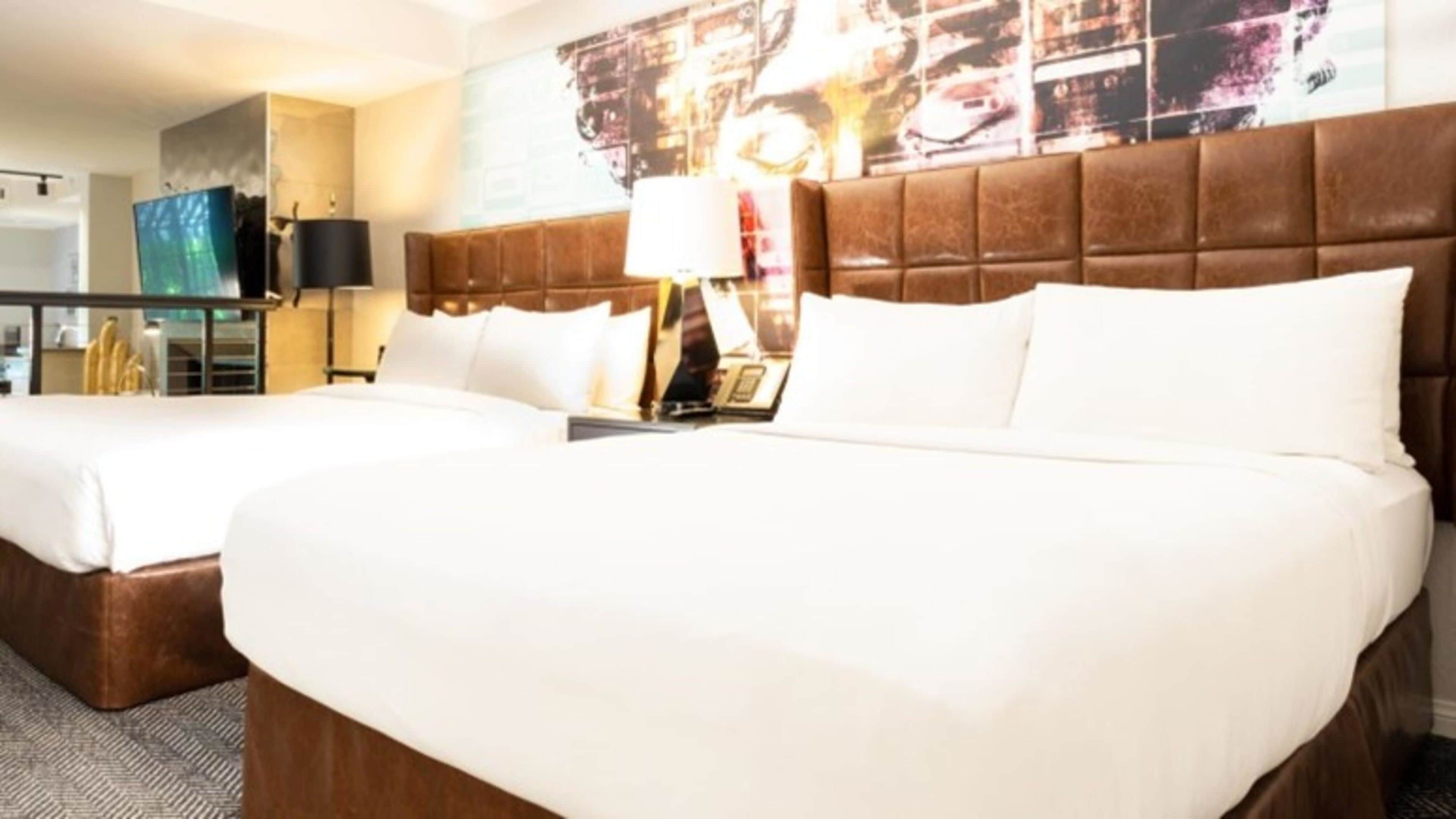 A hotel room with two beds, a brown headboard, a desk with a lamp, and a large abstract painting on the wall.