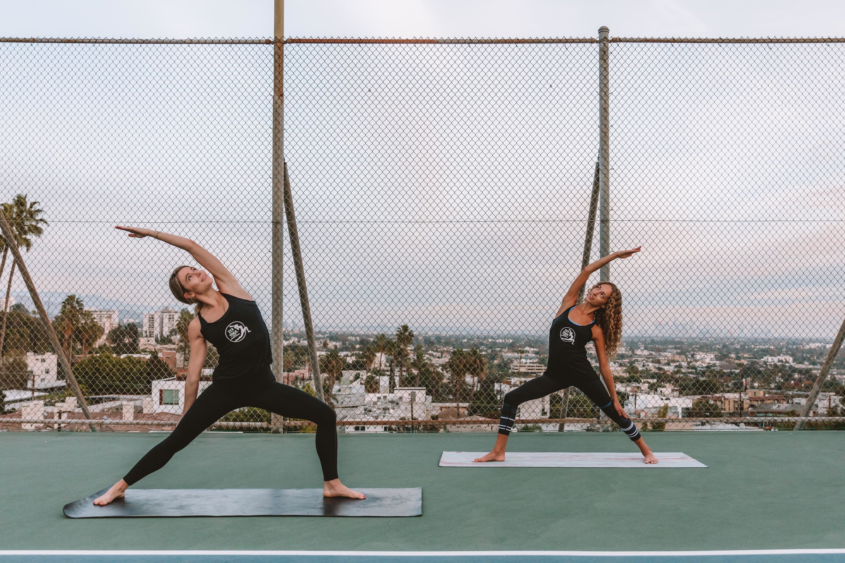 Two individuals practicing yoga on mats on an outdoor court with a cityscape in the background.