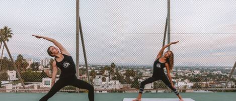 Two individuals practicing yoga on mats on an outdoor court with a cityscape in the background.