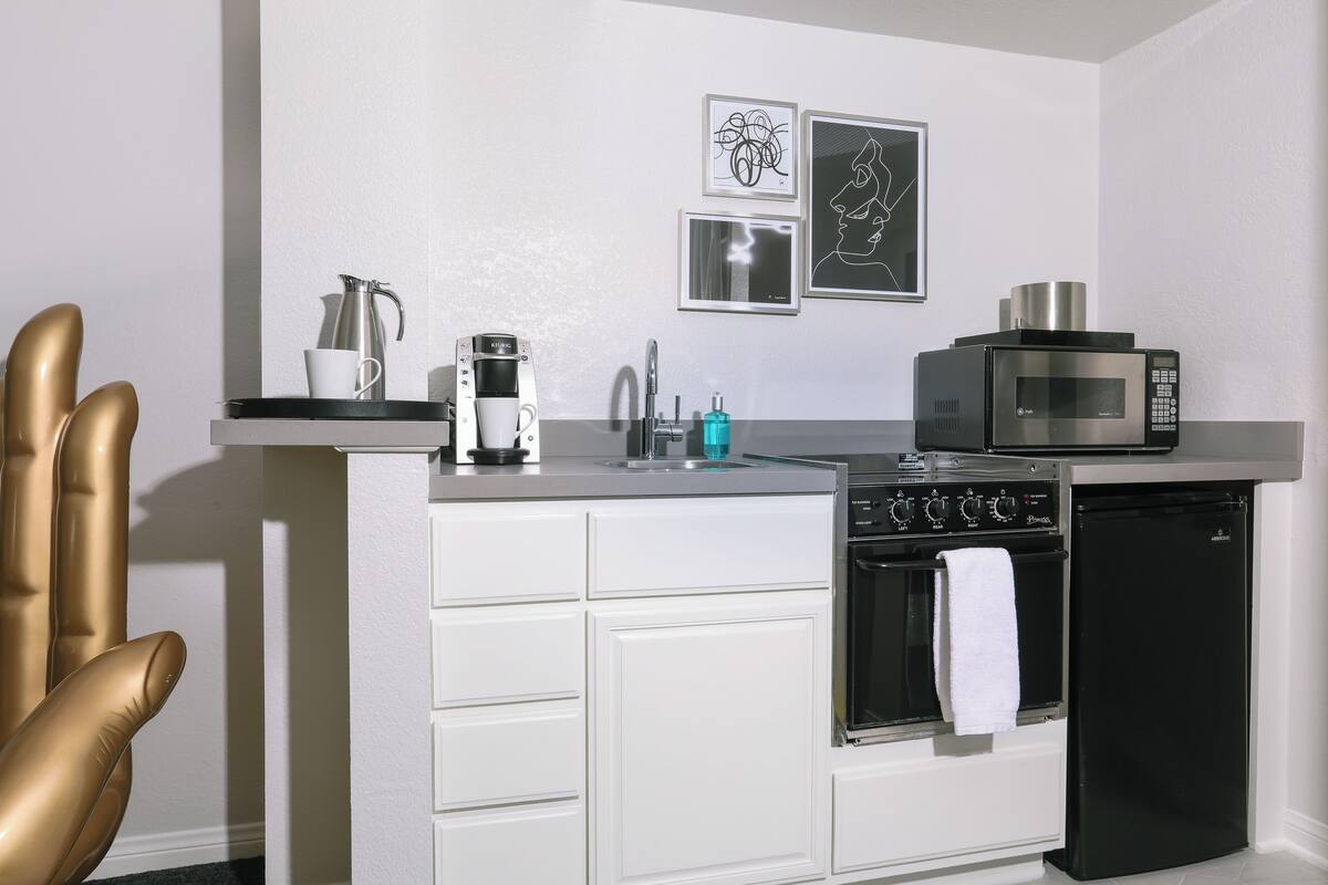 A compact kitchen with a microwave, oven, and coffee maker. There are framed pictures on the wall and a gold chair in the corner.