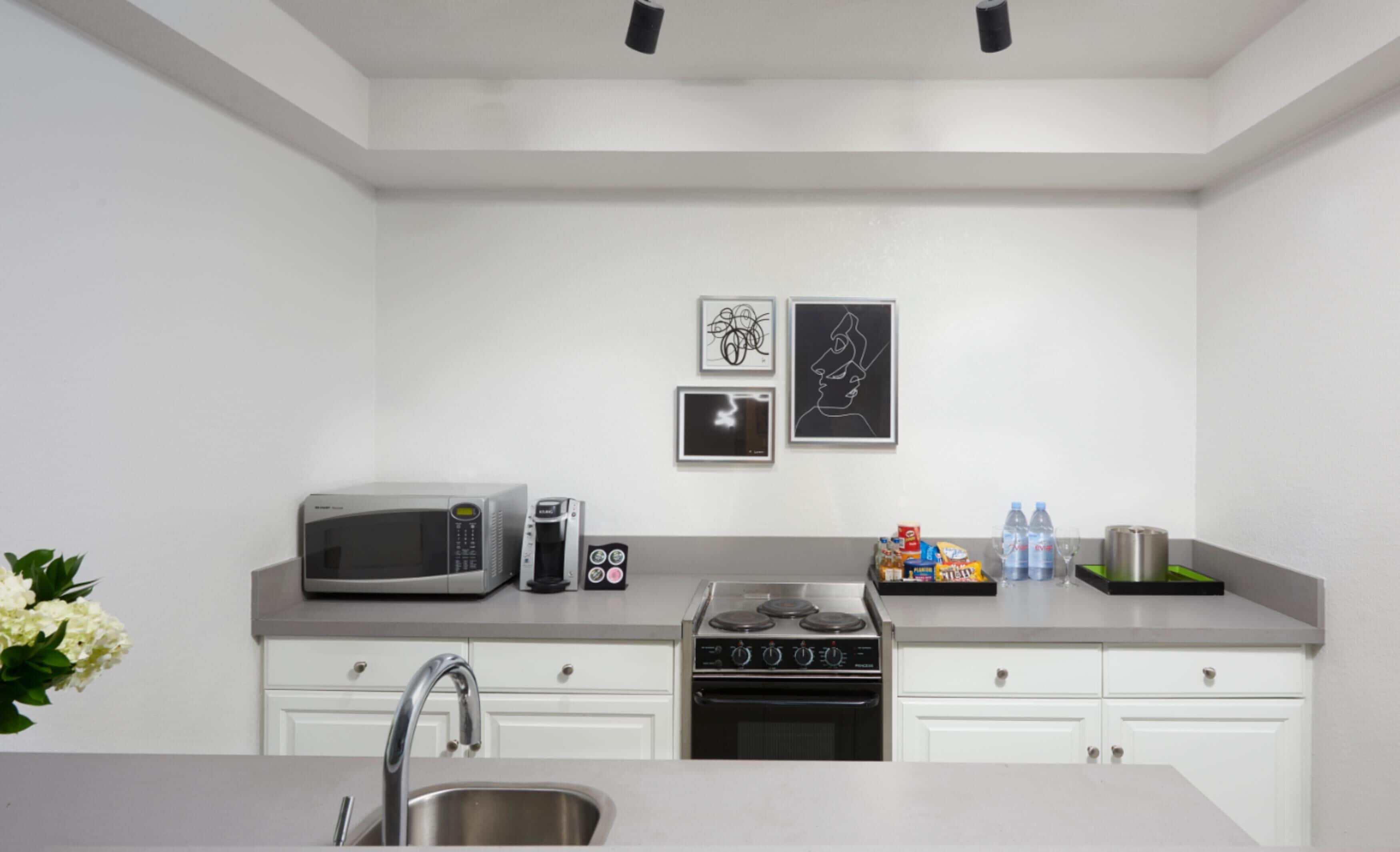A modern kitchen with white cabinets, a stainless steel sink, a microwave, a stove, and a coffee maker. There are framed artworks on the wall and a small selection of snacks and bottled water on the counter.