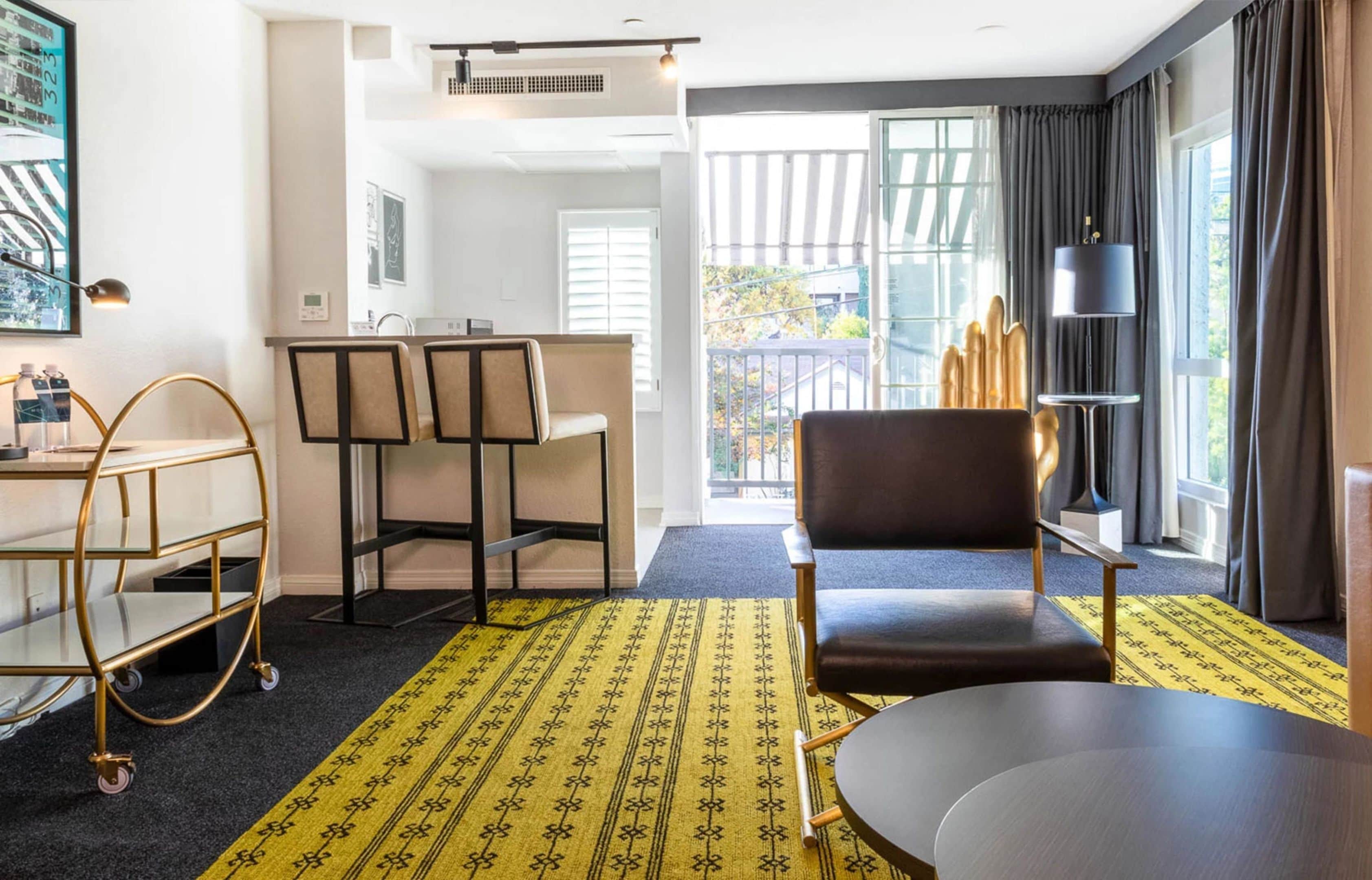 A hotel room with a dining area, a sofa, a table, a cart, and a view of the outside.