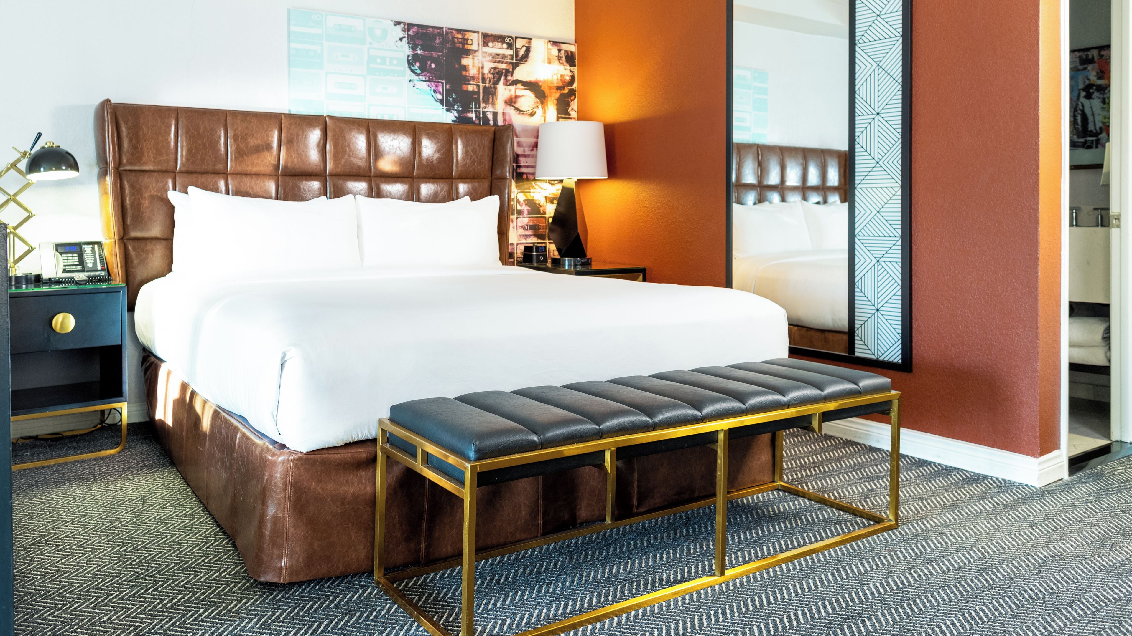 Habitación de hotel con una cama grande, una mesita de noche con lámpara, un banco y una alfombra estampada.