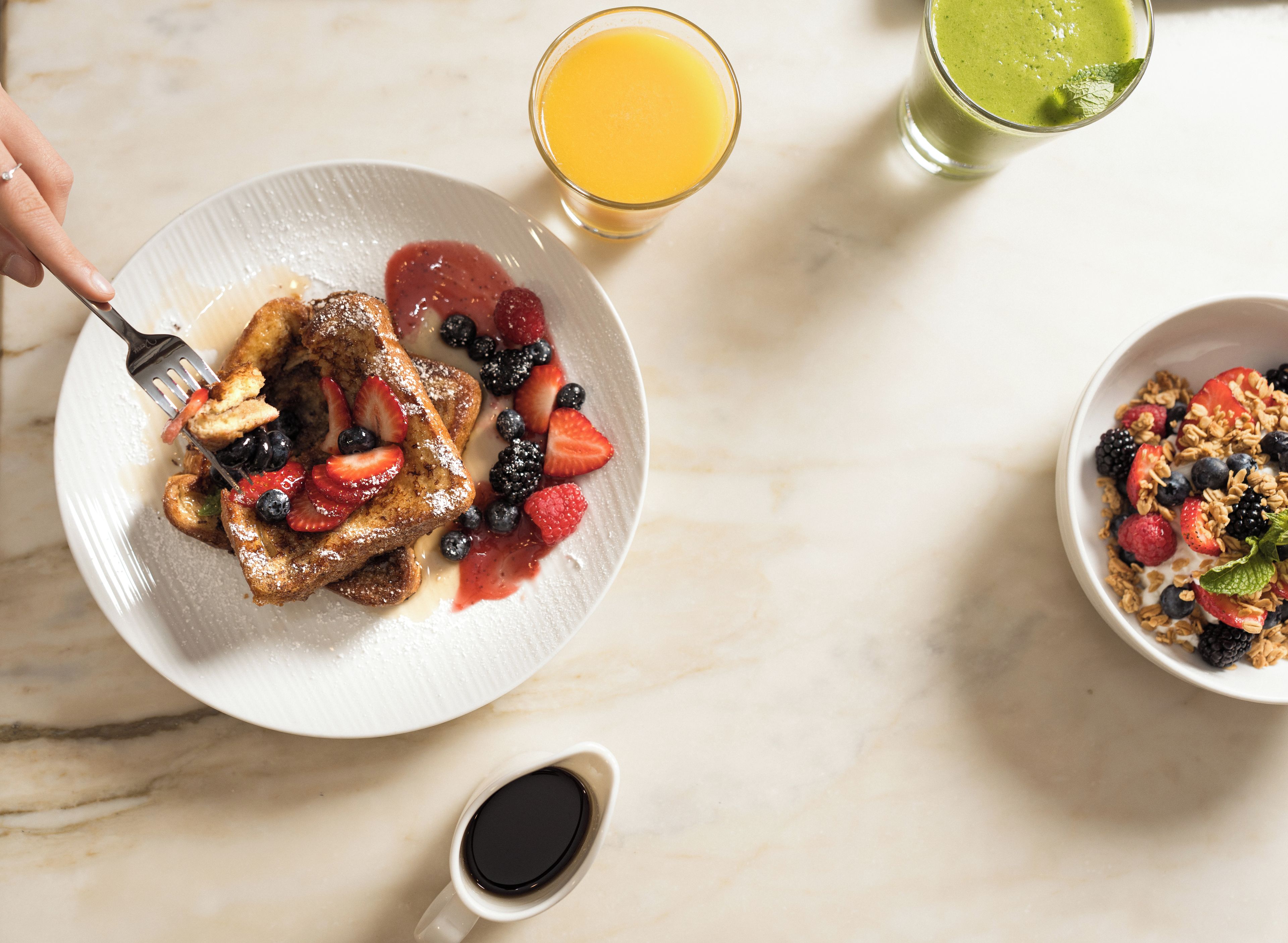 A breakfast setting with a plate of French toast, berries, a glass of orange juice, and a bowl of cereal with fruit.
