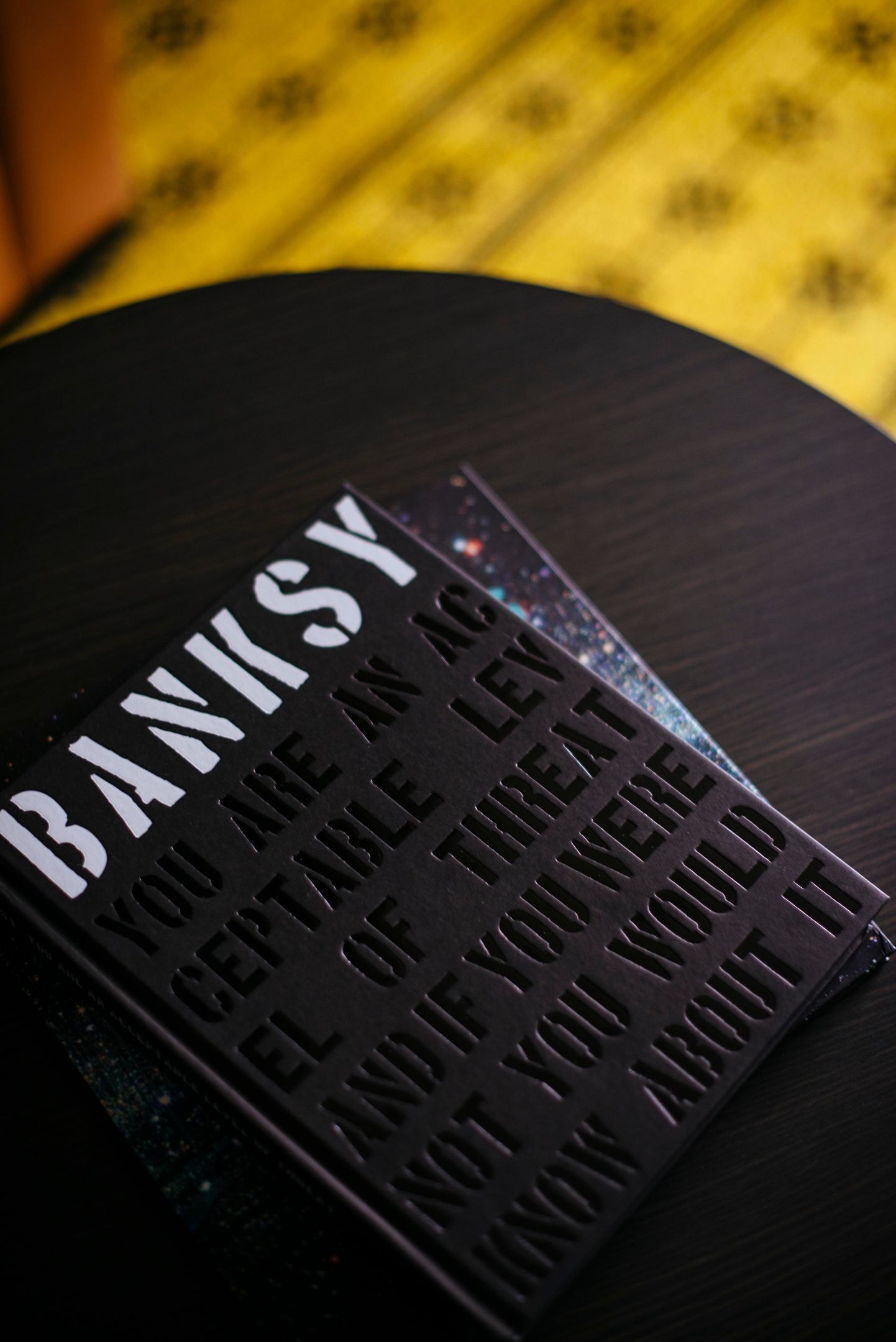 A black surface with embossed text reading 'BANKSY' and 'YOU ARE AN ACCEPTABLE LEVEL OF THREAT IF YOU WERE AND YOU WOULD KNOW ABOUT IT.'