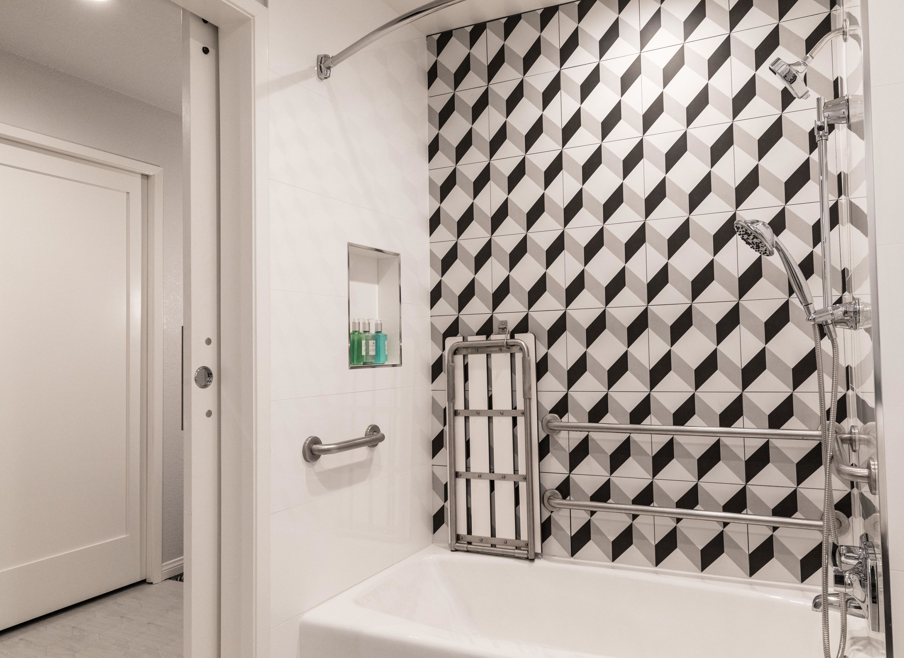 Een badkamer met een wand met een geometrisch patroon, een badkuip, een douche met een handdouche, een handdoekhouder en een klein spiegel met toiletartikelen.