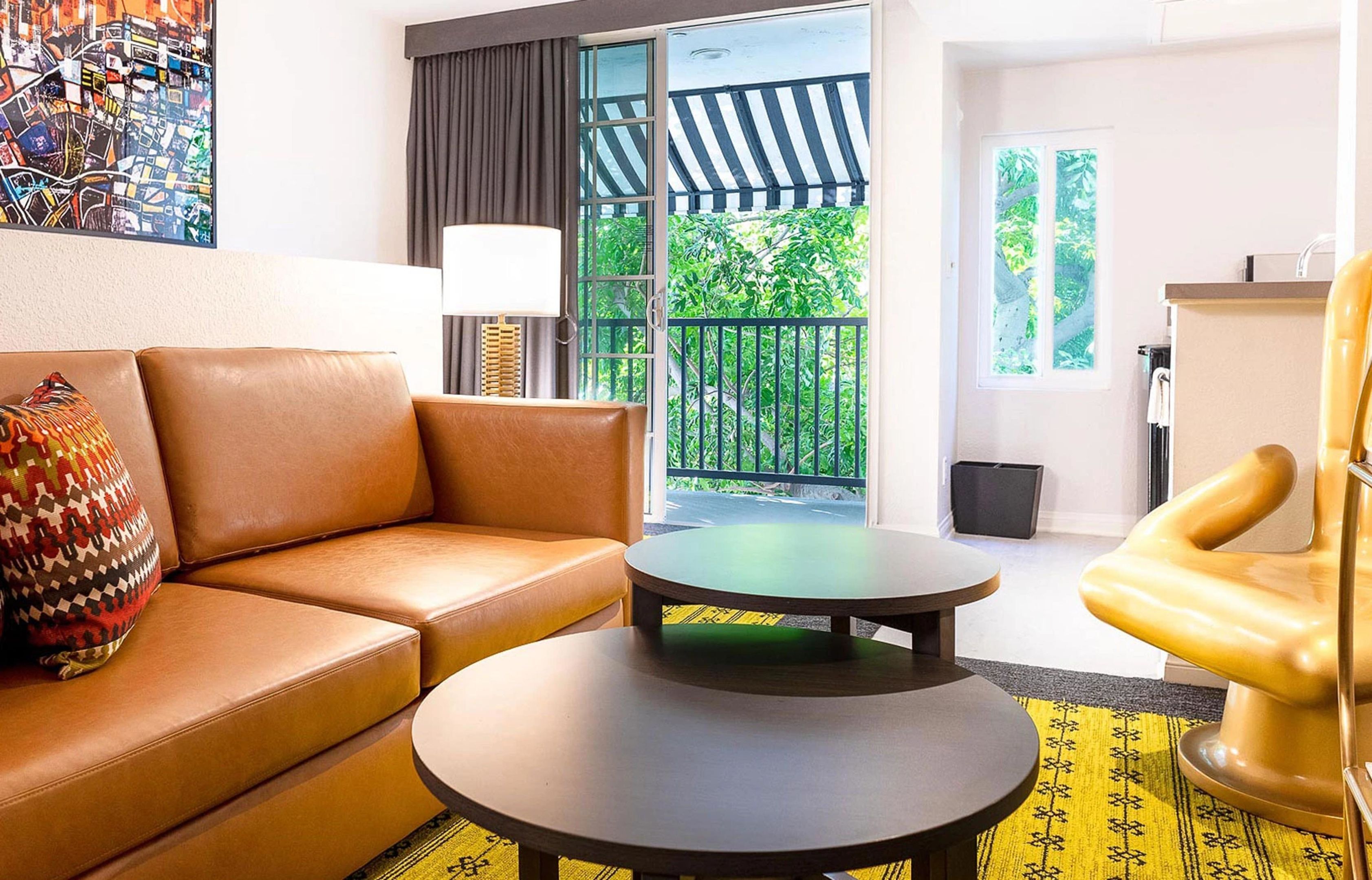 Una habitación con un sofá de cuero marrón, una mesa redonda, un sillón amarillo, un ventanal con vistas a la vegetación y un cuadro abstracto colorido en la pared.