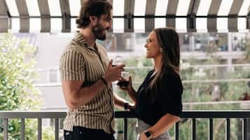 Two people standing on a balcony with a railing, holding glasses.