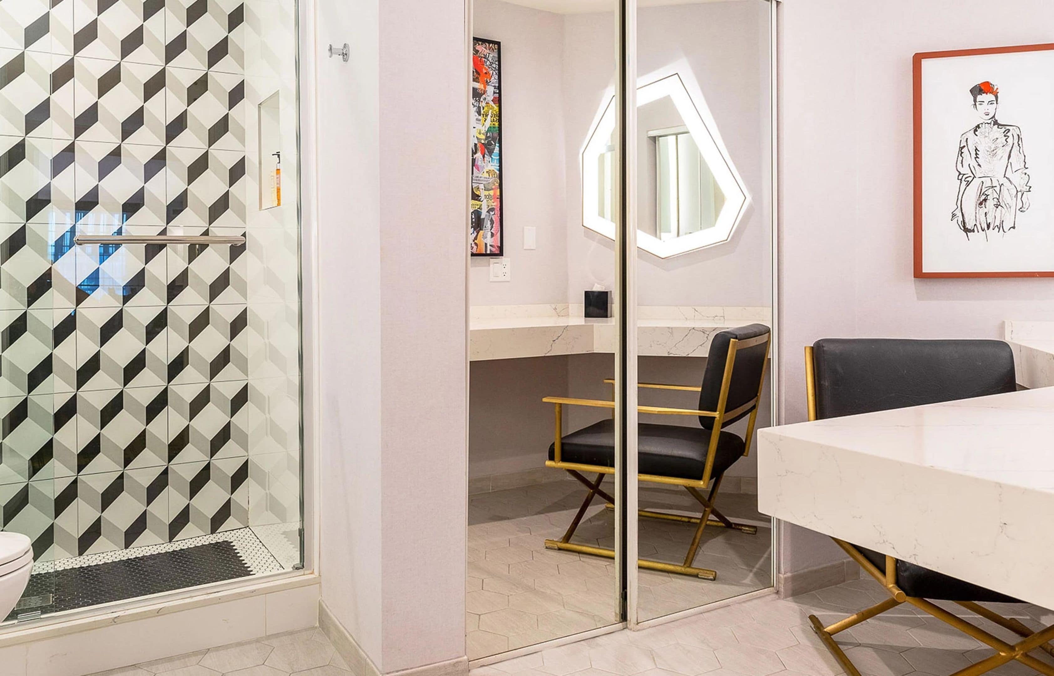 A modern bathroom with a geometric tile backsplash, a glass shower enclosure, a white sink, and a black chair.
