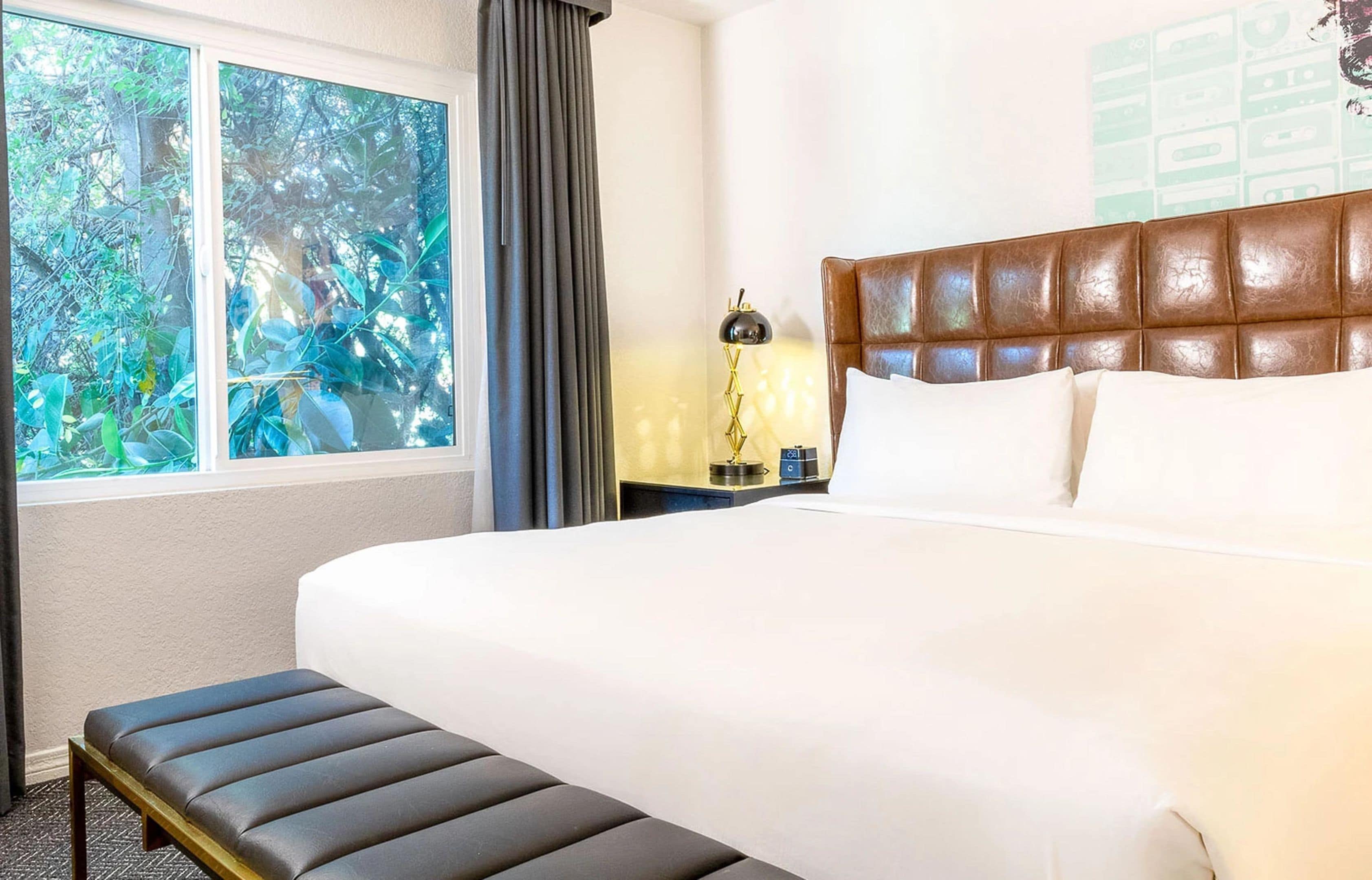A hotel room with a large bed, a bench, a nightstand with a lamp, and a window with a view of trees.