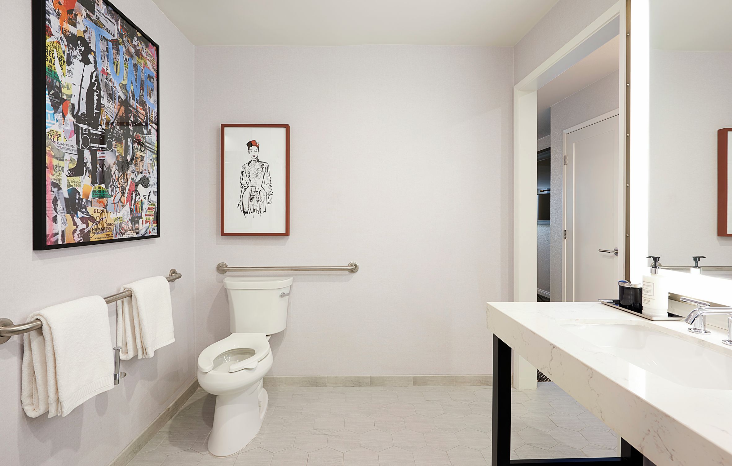 Baño con inodoro, barra de apoyo, lavamanos y espejo. Hay un cuadro en la pared.