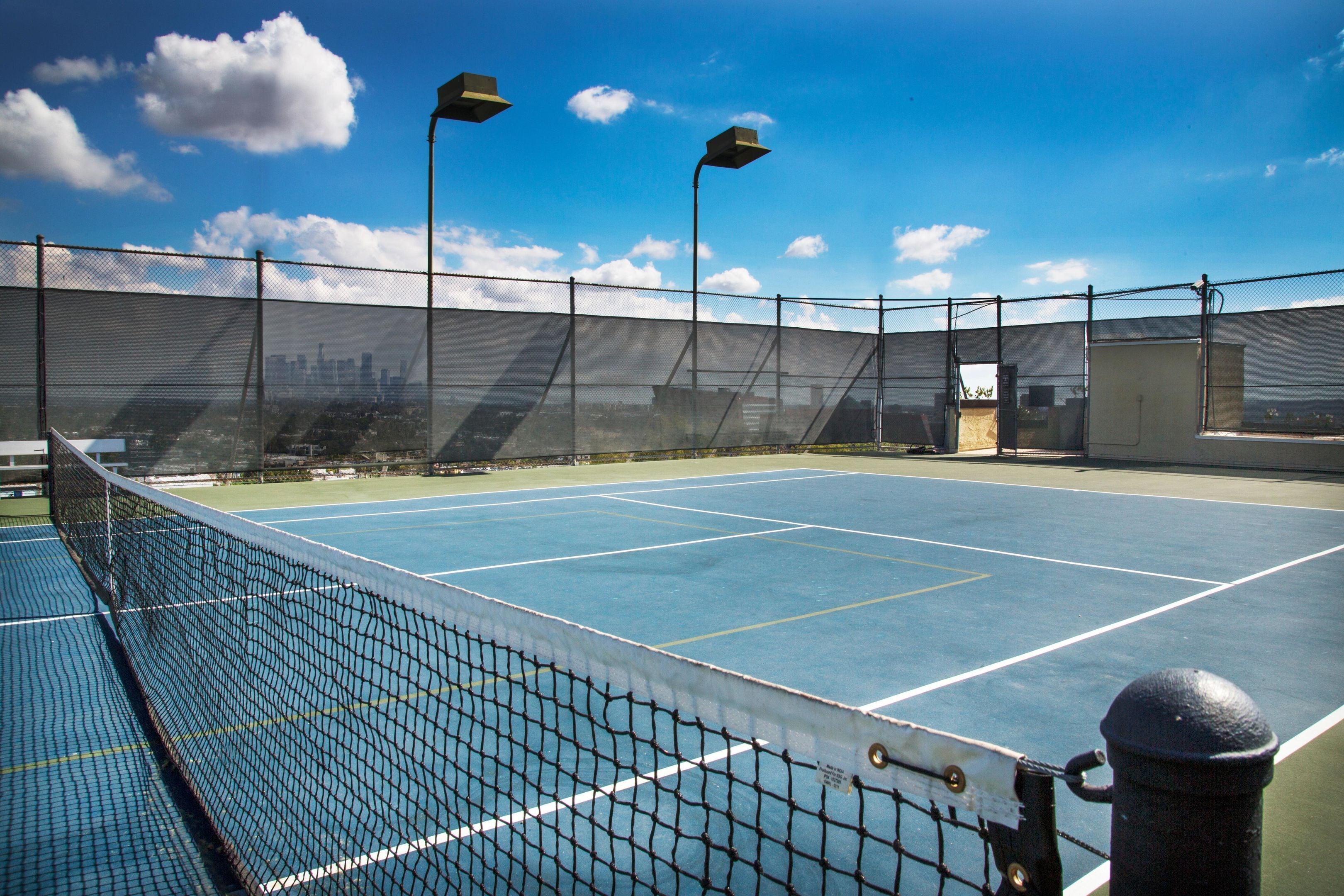 A tennis court with a net, surrounded by a fence, and two street lamps.