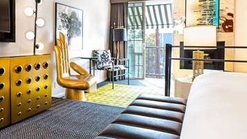 Een hotelkamer met een groot bed, een gouden fauteuil, een bijzettafeltje, een vloerlamp en een groot raam met gordijnen.